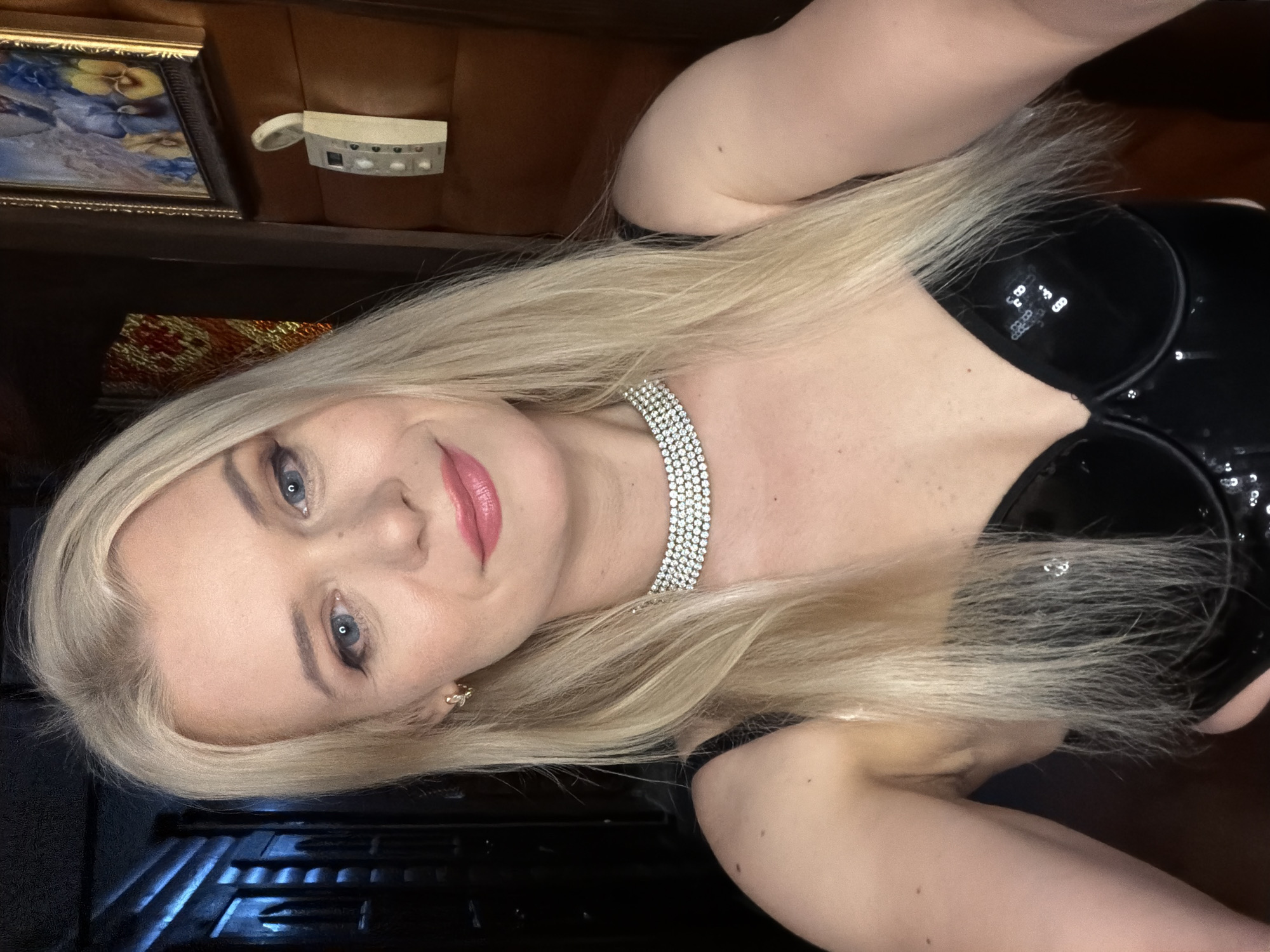 The media file is a close-up, portrait-style photograph of a blonde woman with blue eyes and pink lipstick. She is wearing a sparkly choker necklace and a black, shiny top. The image is taken indoors, with a dimly lit background that suggests a domestic or private setting. To the left of the woman, a framed painting with floral imagery is visible. Further to the left, a small white object with buttons is attached to a brown surface, possibly a wall or furniture. The lighting is low, and the overall mood appears to be intimate or sensual. No specific time of day or weather conditions can be inferred. No text is visible in the image. The provided location context is Sochi, Russia.