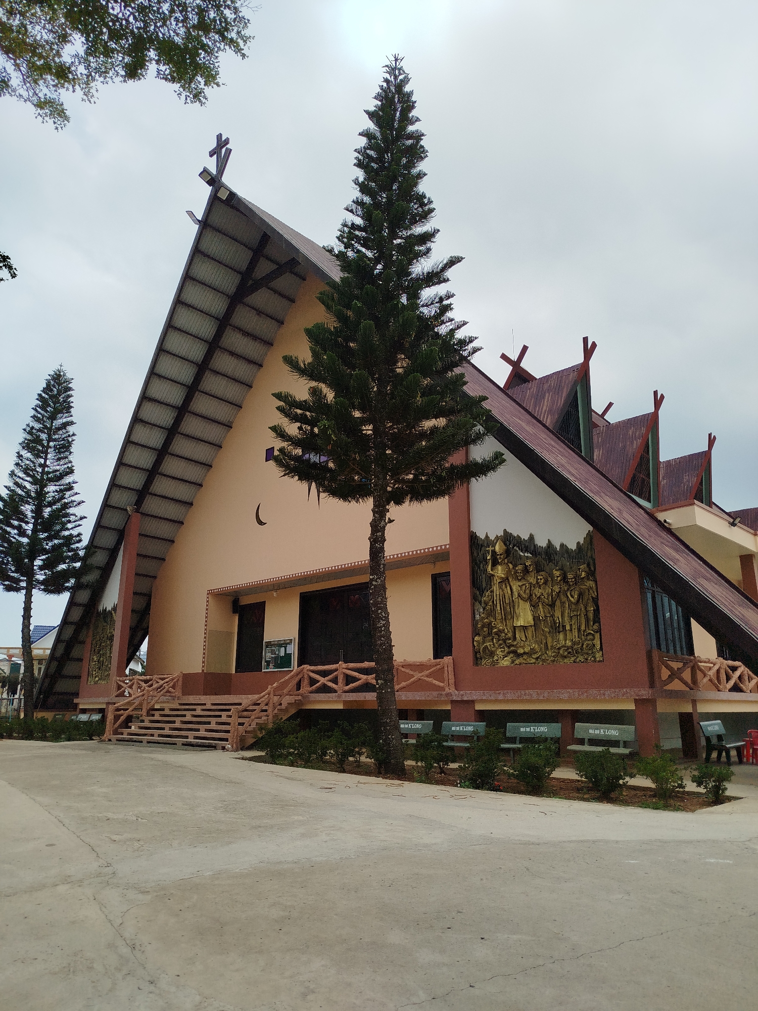 The image shows a church located in K'Long, Vietnam. The building features a steep, metal roof with a cross at its apex. The facade is painted in a pale beige color, accented with darker brown trim. A large bas-relief sculpture is installed on the right side of the building. A crescent moon shape is visible near the center of the front facade. Two tall, thin evergreen trees flank the building. Benches are positioned along the front of the church, marked with the name "Nha tho K'Long". The sky is overcast. The ground is paved with concrete. Steps lead up to the main entrance of the church. A wooden railing borders the steps and the raised platform in front of the building.