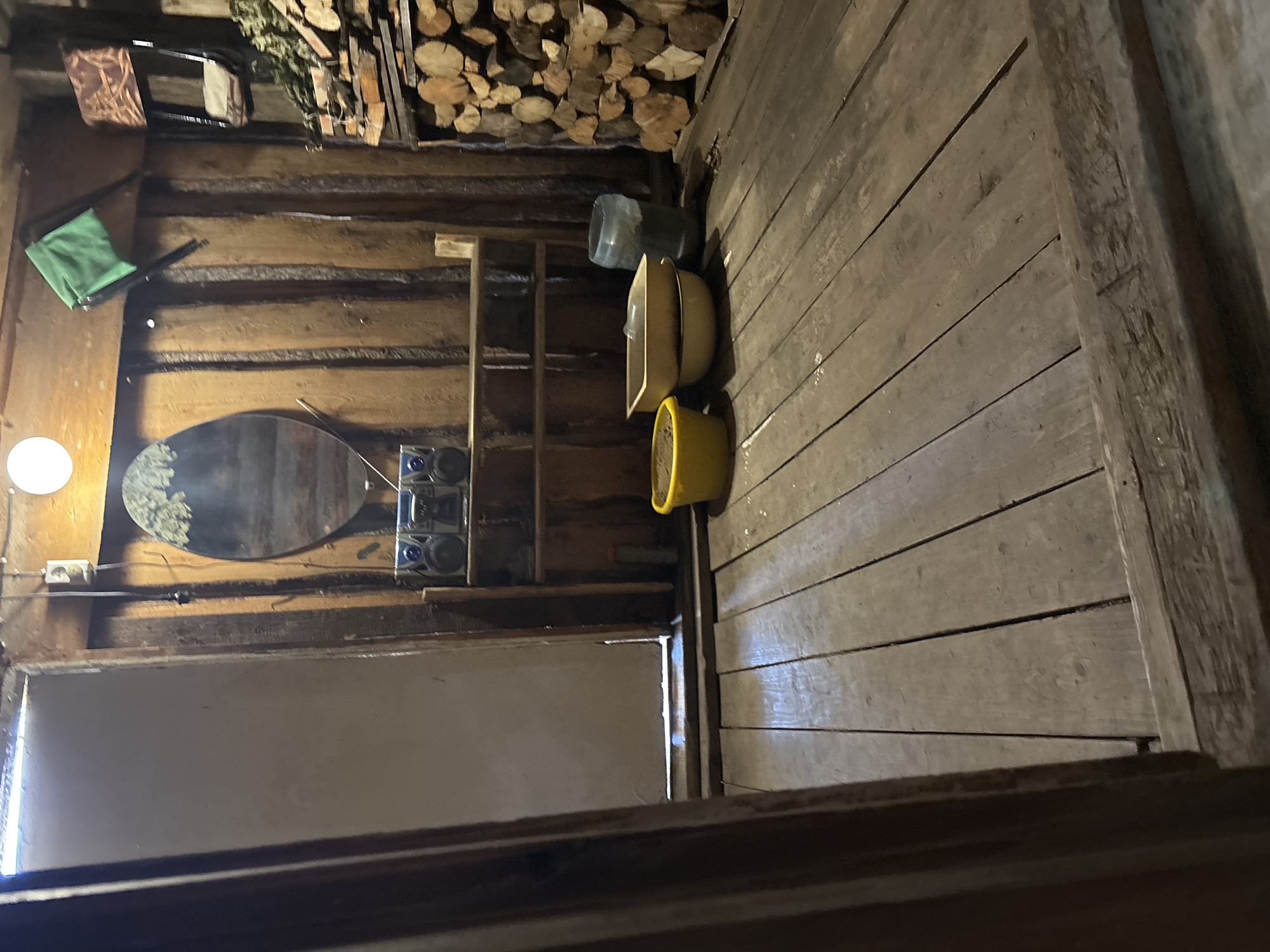 This image depicts the interior of what appears to be a rustic wooden structure, potentially located in Ферма, Russia given the provided location. The walls and floor are constructed of unfinished wooden planks.
Along one wall are arranged several items including firewood stacked high, a green cloth item, and a mirror. A set of audio speakers is visible along with shelving containing other miscellaneous objects. A light fixture emits a soft glow near the mirror.
On the floor, there are plastic containers, including buckets. The image is taken from an elevated perspective, looking down into the scene.