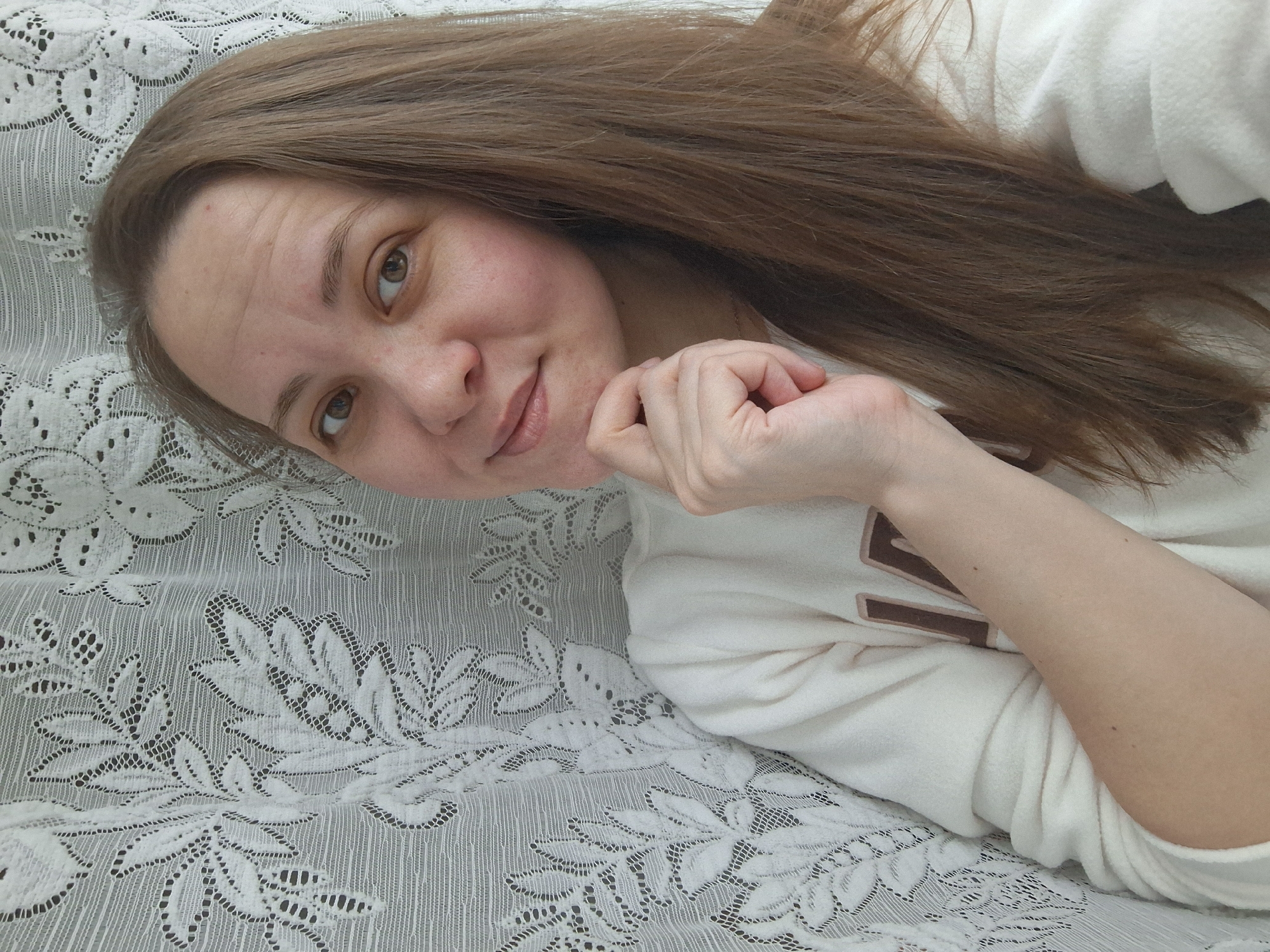 A woman with brown hair is lying on a white lace fabric. She is wearing a white sweatshirt with brown lettering on the chest. Her left hand is resting on her chin, and she is looking directly at the camera with a slight smile. The background consists of the white lace fabric. The image was taken in Chaikovsky, Russia.
