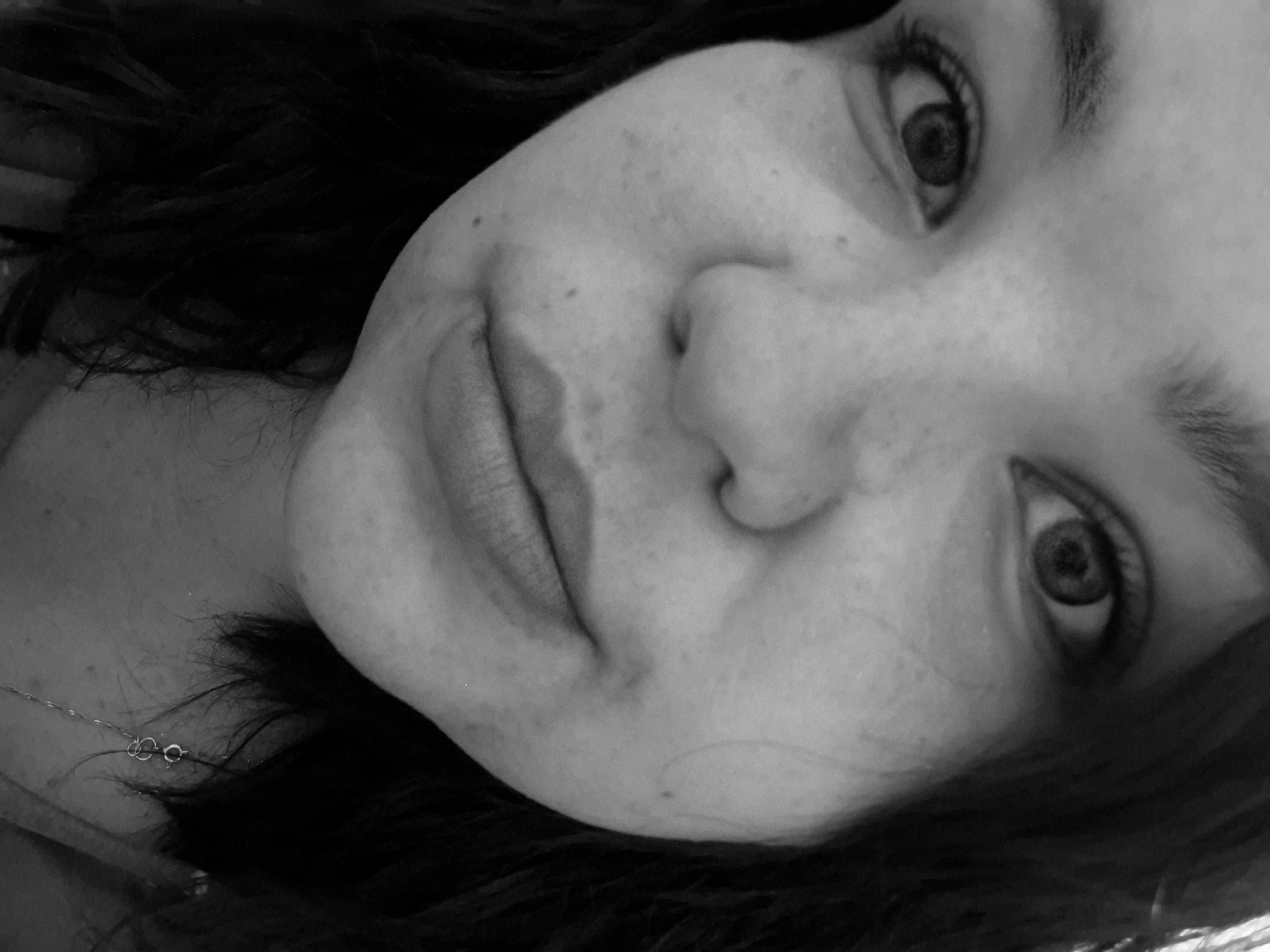 A close-up, black and white portrait shows a person's face at an angle, filling most of the frame. The subject's eyes are open and looking slightly upward. The nose is prominent, and the lips are closed with a slight suggestion of a smile. Freckles or blemishes are visible on the skin. Dark hair frames the face. A delicate necklace is visible at the lower left of the image. The scene's location in the city of Марковский, Russia cannot be determined from the image.
