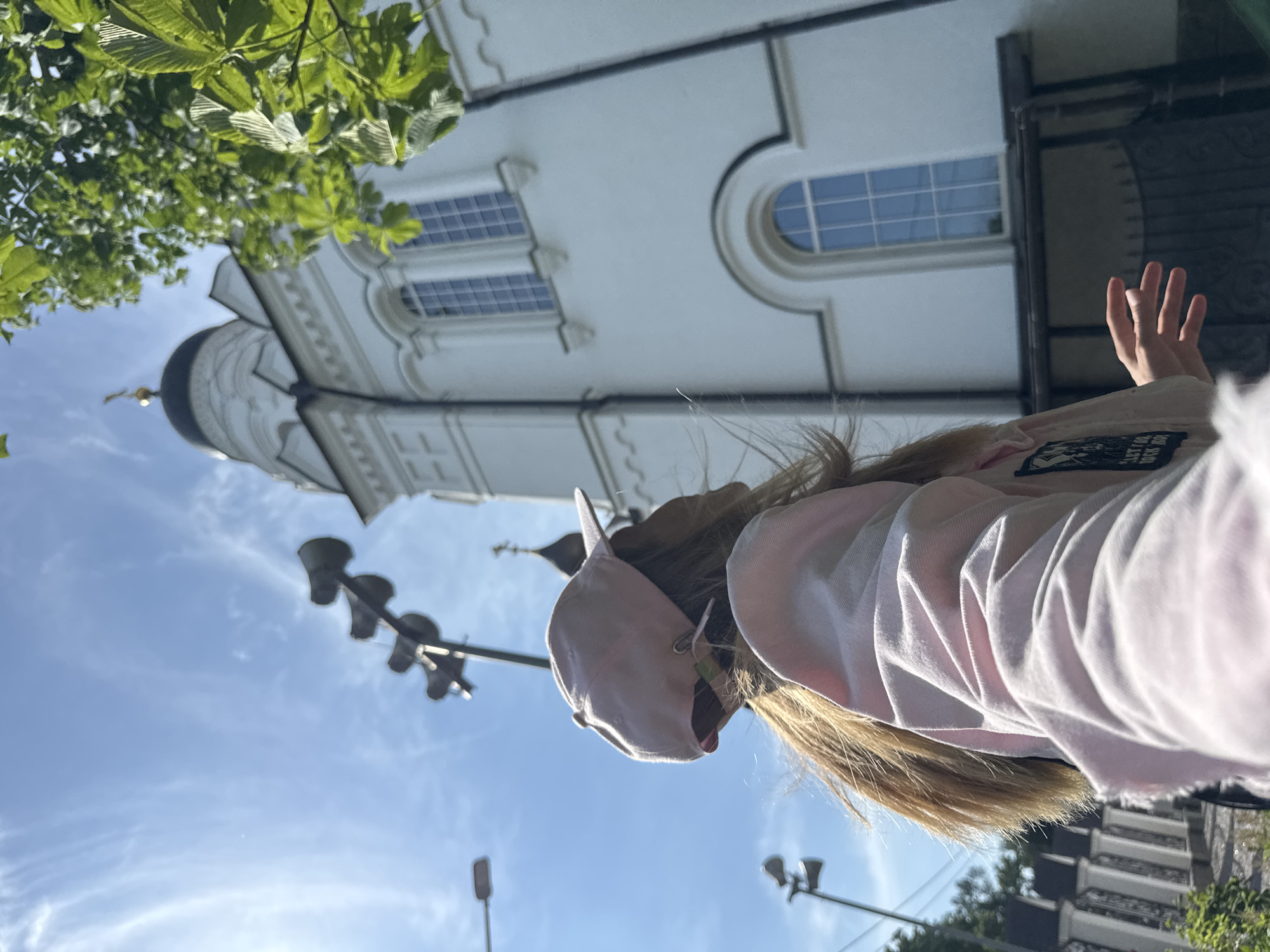 The image shows a person from behind, wearing a pink hoodie and a pink baseball cap. The person's blonde hair is visible. They are raising their right hand, fingers spread, towards the sky. In the background, there is a white building with a black dome, possibly a church or cathedral, with arched windows. Green leaves from a tree are in the upper left corner of the frame. The sky is blue with wispy clouds. Several streetlights are visible in the lower part of the image, suggesting an outdoor setting. The lighting indicates it is daytime, likely morning or afternoon, with bright sunlight casting shadows. There is a patch on the hoodie with text that is partially visible and difficult to read, but appears to say "FOR THEM". The image appears to be taken from a low angle, looking upwards. The scene is likely a public space in Lomonosov, Russia, given the architectural style of the building and the presence of streetlights. The person's gesture might suggest a greeting, a farewell, or a general expression of optimism or joy.