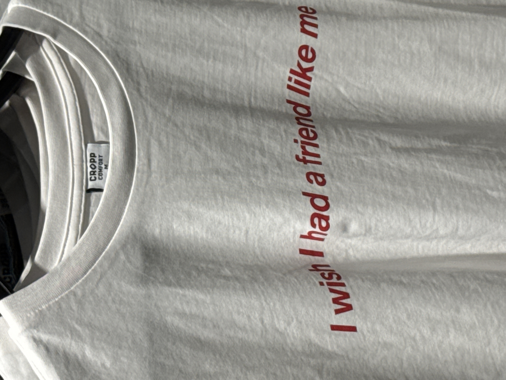 The image shows a light-colored t-shirt, likely white or off-white, displayed vertically. The collar of the t-shirt is visible on the left side of the frame, revealing a tag. The main text on the t-shirt, printed in red and slanted, reads: "I wish I had a friend like me".
The tag attached near the neckline features the brand name "CROPP" in bold capital letters, with "COMFORT" printed below it, and the size "M" (for Medium) beneath that. The t-shirt fabric shows some subtle texture and gentle folds, suggesting it is either hanging or lightly creased. The background is softly blurred, hinting at additional clothing items or a retail environment, as might be found in a store in Sofia, Bulgaria. The lighting is diffused and even, providing a clear view of the garment and its text. No people are visible, and no specific time of day or weather conditions can be discerned.