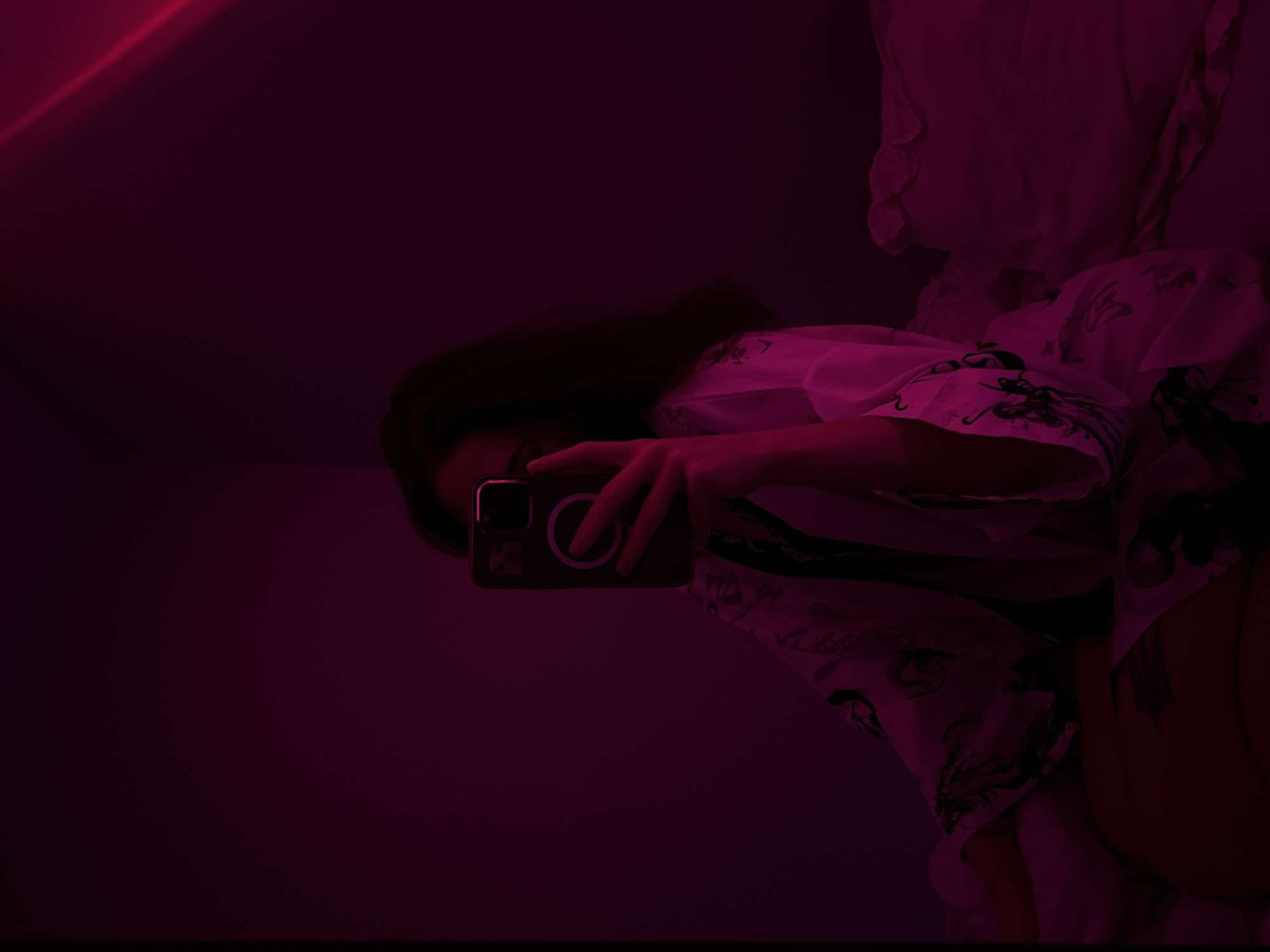 A person is taking a selfie in a room with pinkish-purple lighting. The person is holding a smartphone with a dark-colored case and a white circular magnet on the back. They are wearing a white robe with black intricate designs. The room appears to be a bedroom, with a bed visible in the background, covered in white bedding. The overall mood of the image is dimly lit and atmospheric. There is no information or visual cues in the image that suggest this scene is located in Ternopil, Ukraine.