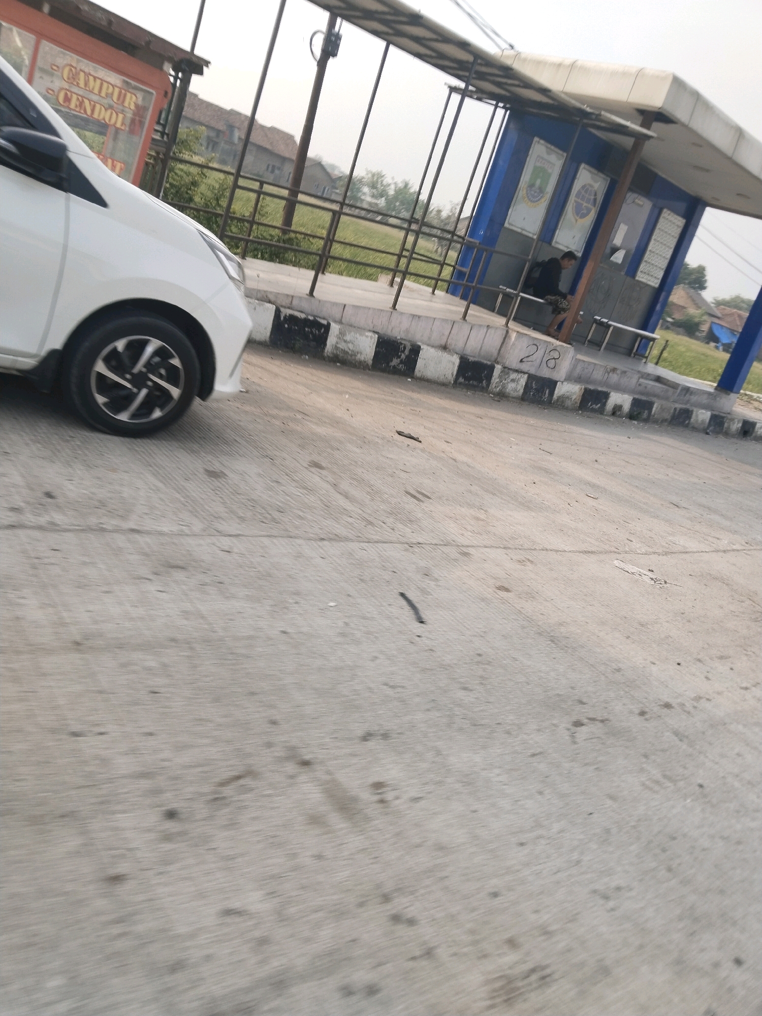 The image depicts a street scene in Serang, Indonesia, likely near a bus stop or a local establishment. In the foreground, the left side of a white car is visible, with its front wheel and part of the bumper in focus. The car appears to be stationary.
To the right, a bus stop structure is partially visible. It has a blue and white color scheme and is supported by metal poles. A person is seated on a bench inside the bus stop, facing away from the camera and towards the interior of the structure. The person is wearing a dark top and patterned shorts. The bus stop has some signage displayed on its walls, one of which appears to be a circular emblem. Below the bus stop, on a concrete barrier, the number "218" is spray-painted in black.
Behind the bus stop and the car, a fence runs along the side of the road, beyond which are some buildings with tiled roofs and trees. The overall environment appears to be an outdoor setting, possibly a roadside area in a moderately developed part of Serang. The road surface is concrete, with visible lines and some scattered debris. The sky is overcast, suggesting a cloudy day. To the left of the car, there is an orange structure with text that is partially legible, possibly indicating a food or drink stall with the words "CAMPUR CENDOL" visible.