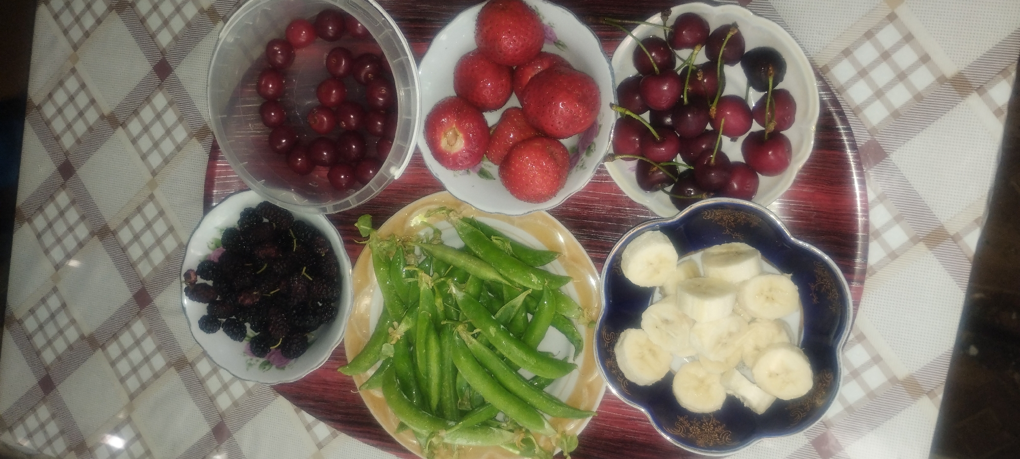 A top-down, high-angle shot captures a collection of fresh fruits and vegetables arranged on a checkered tablecloth. The setting appears to be indoors, likely a kitchen or dining area. The fruits include mulberries, cherries, strawberries, and sliced bananas, all presented in separate bowls or containers. Additionally, a plate of green peas in their pods is centrally located. The overall scene is one of abundance and healthy eating. The image was taken in Zaporizhzhia, Ukraine.