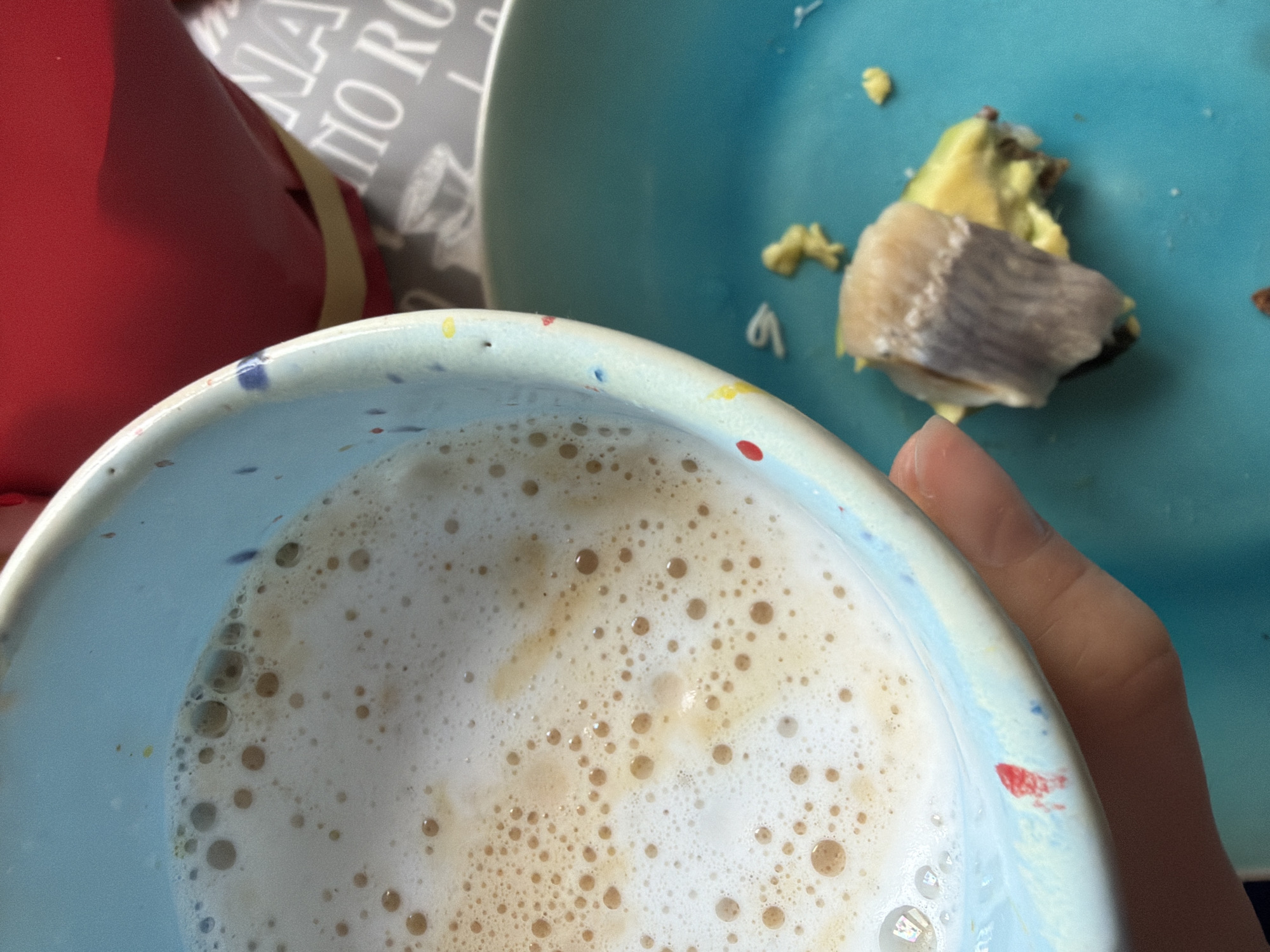 A high-angle, close-up shot features a light-blue speckled cup filled with a foamy beverage, possibly a latte or cappuccino. To the right of the cup, a turquoise plate holds a small portion of food, seemingly avocado and a slice of fish. A hand is partially visible, holding the cup. In the background, there's a red object and a gray patterned surface. The location is Unknown, Unknown. No people are fully visible in the image.