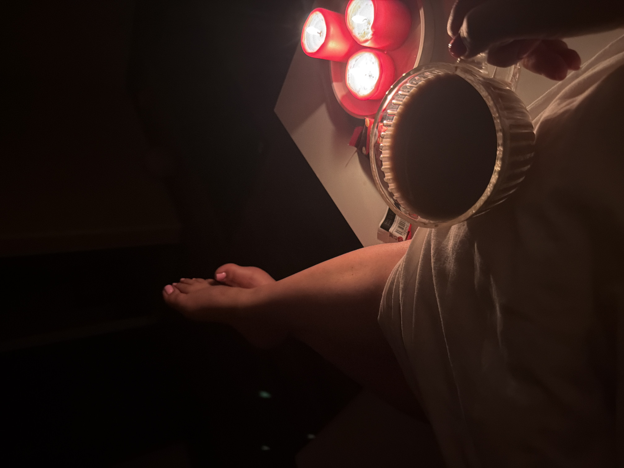 The image shows a person sitting in a dimly lit room, holding a glass of dark liquid. The person's left leg is visible, with pink nail polish on the toes. A set of three red candles illuminates the scene from a nearby surface. The location and city of Leningradskaya, Russia are not discernible from the provided image.