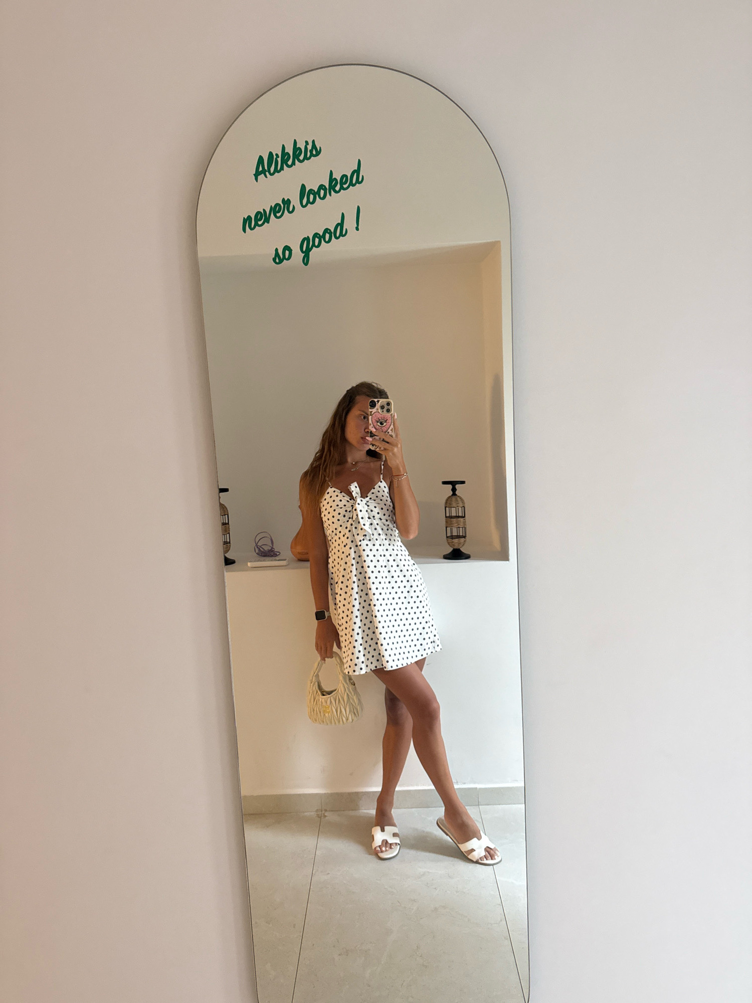 A full-length mirror with an arched top leans against a light pink wall. Green text on the mirror reads "Alikkis never looked so good!". A young woman with long brown hair is reflected in the mirror, taking a selfie with her phone. She is wearing a white polka dot mini dress with a bow detail at the bust, a beige quilted handbag, and white slide sandals. She is standing in what appears to be a room with white walls and tiled floors, likely in Stavros, Greece, as suggested by the text on the mirror. A shelf with decorative objects is visible behind her reflection.
