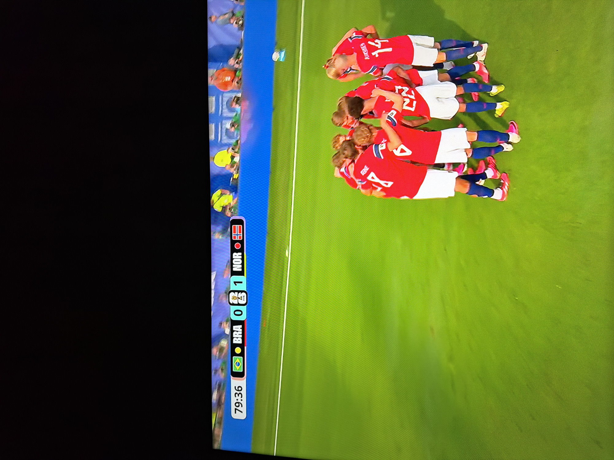 The image captures a television screen displaying a soccer match. A scoreboard at the top shows the time as 79:36, with Brazil (BRA) at 0 goals and Norway (NOR) at 1 goal. The players, all wearing red jerseys and white shorts, are huddled together in the center of the green soccer field, appearing to be celebrating or strategizing. Their numbers are visible on their backs, including 8, 6, 22, and 14. The field has a white line marking its boundary. In the background, spectators are visible in the stands. The image does not provide any information regarding the city of Ognianovo, Bulgaria.