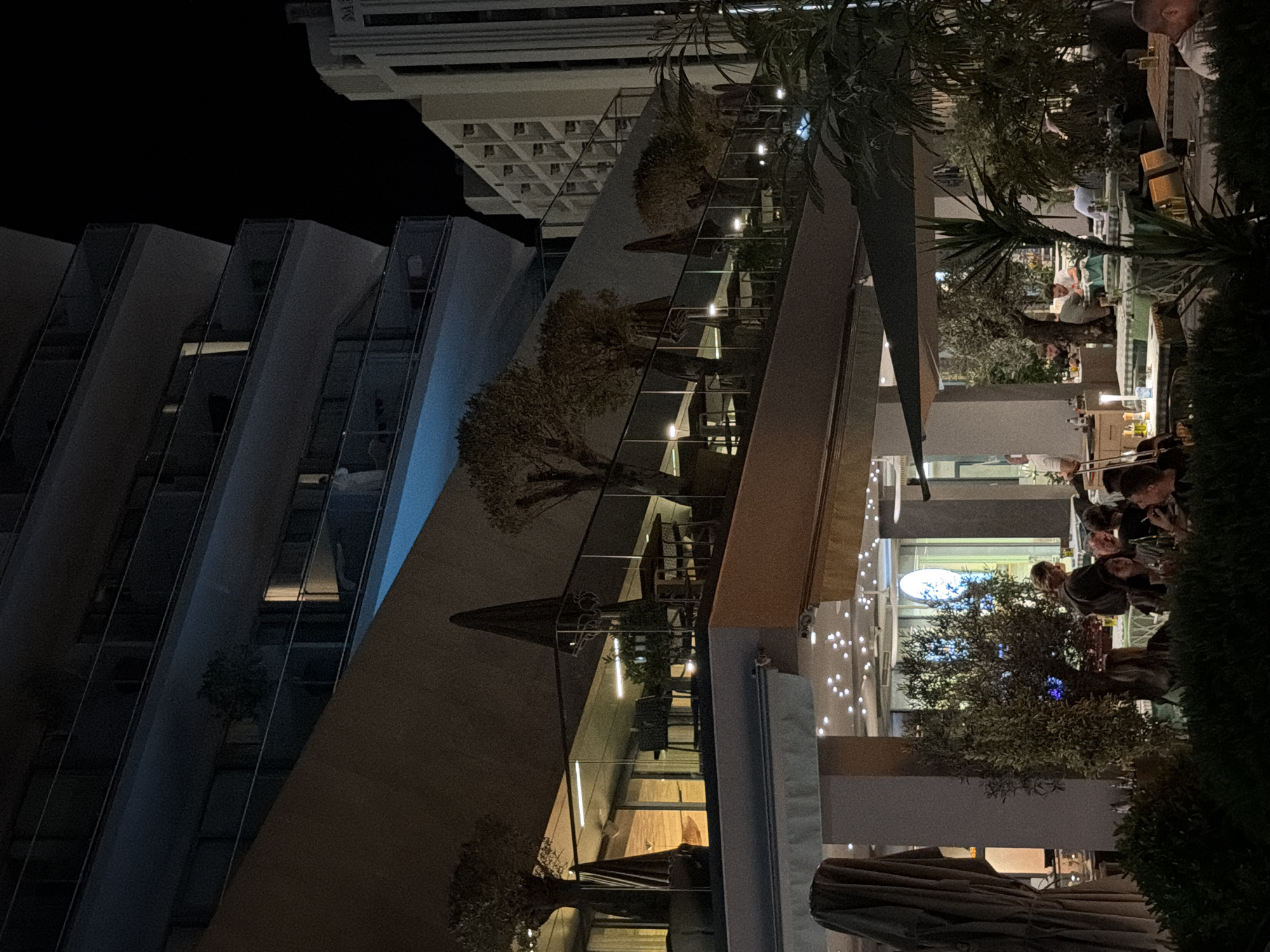 At night in Rafailovići, Montenegro, an outdoor restaurant seating area is visible. The scene is illuminated by artificial lights and the ambient glow from nearby buildings. In the foreground, patrons are seated at tables, some engaged in conversation, others with drinks in hand. To the left, large closed umbrellas are stacked. Beyond the dining area, the facade of a multi-story building, likely a hotel or apartment complex, curves upwards and is partially obscured by a slanted, lighter-colored wall. Smaller trees with sparse foliage are planted on terraces of this building. To the right, another building with a grid-like facade stands against the dark night sky. The overall impression is a lively urban night scene.