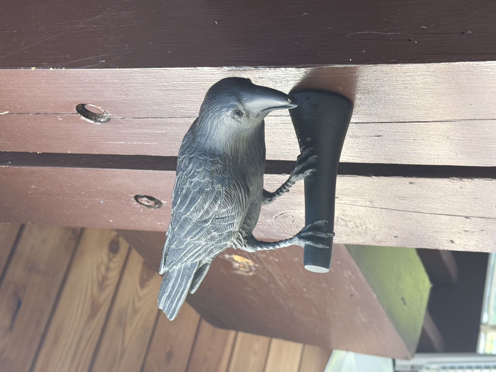 A dark gray, ornamental bird figurine, possibly a crow or raven, is mounted on a dark brown wooden post. The bird is positioned facing towards the right of the frame, with its beak lowered as if pecking at something. Its wings are detailed with a feathered texture. The bird is perched on a dark, cylindrical object attached to the post. The post itself has visible wood grain and appears to be part of an outdoor structure, possibly a porch or fence. In the background, there is a lighter brown wooden ceiling or overhang. The image appears to be taken outdoors, with natural light illuminating the scene. The overall setting does not provide any specific indicators for location within Nizhnyaya Shilovka, Russia.