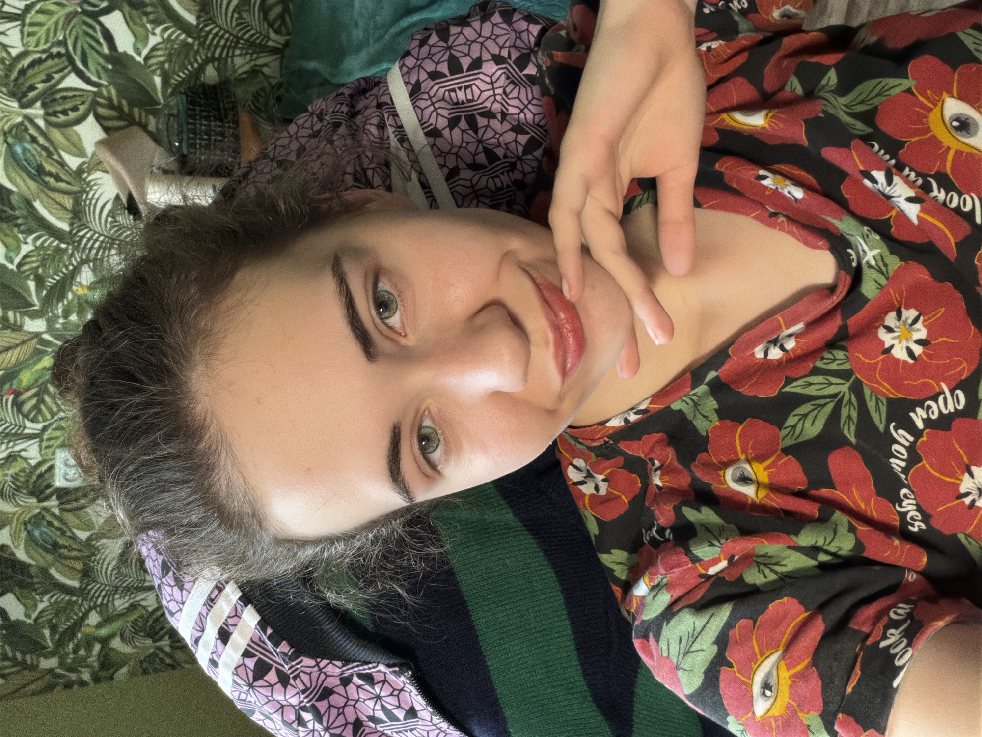 A young woman with dark hair pulled back and green eyes is looking at the camera. She is wearing a black shirt with a red floral pattern and text. Her right hand is resting on her chin. The background consists of a patterned wallpaper with green leaves and birds, and some pillows. The image appears to be a selfie taken indoors. There is no discernible information in the image that would indicate a location in Zavodoukovsk, Russia.