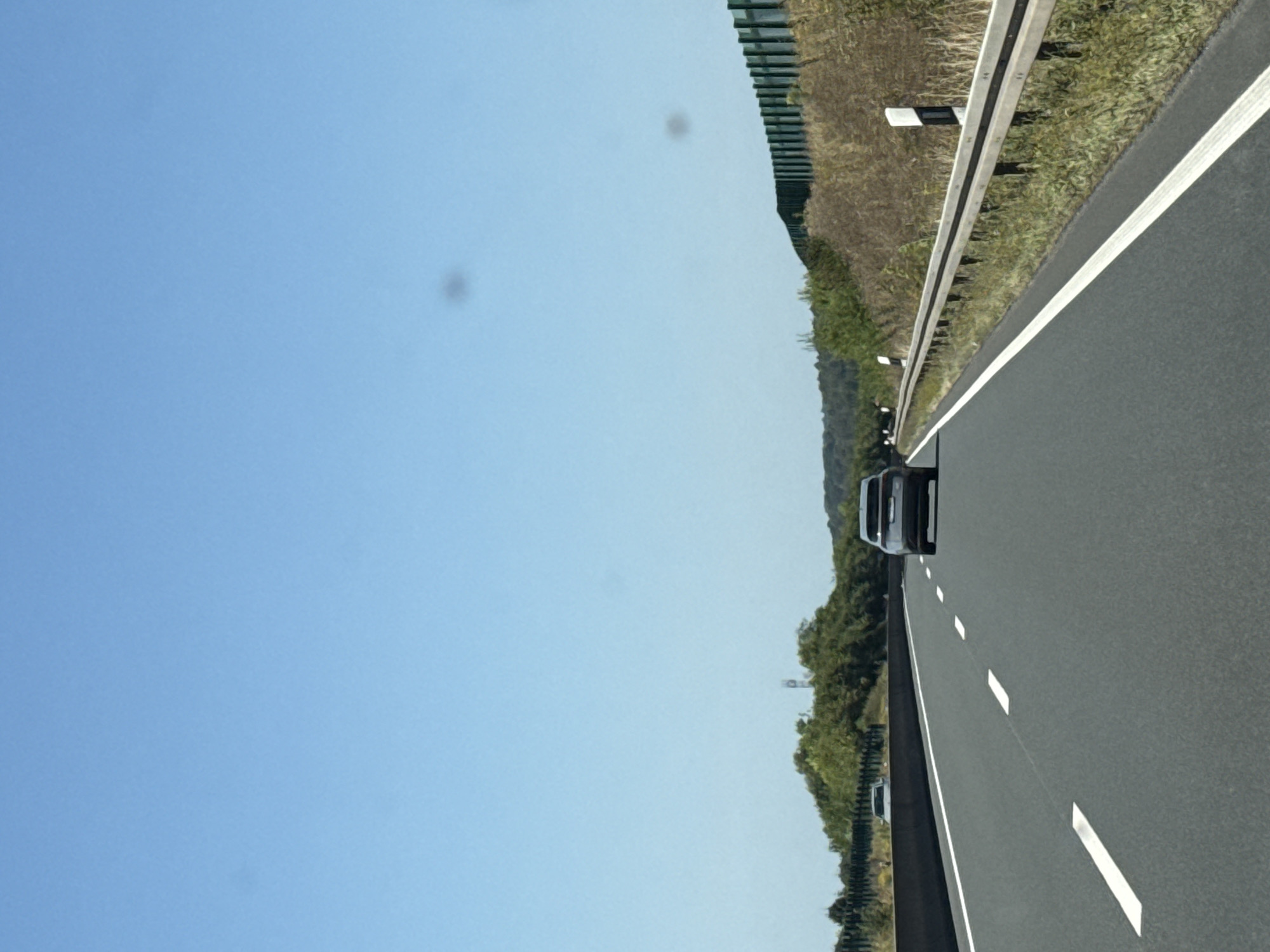 The image shows a view from a car driving on a highway. The highway has multiple lanes and is marked with white lines. On the right side of the highway, there is a metal guardrail and a grassy embankment with some dry vegetation. A green fence is visible further up the embankment. On the left side of the highway, there is a concrete barrier and trees and a hill in the distance. The sky is clear and blue. A dark-colored car is driving in the right lane ahead of the camera car, and another vehicle is visible in the left lane in the distance. Two dark spots are present in the sky, likely lens artifacts. The scene appears to be in a rural or suburban area given the presence of trees and hills. The location in the city of Bad Sachsa, Germany is not discernible from the image itself, but the highway infrastructure is consistent with German autobahns.