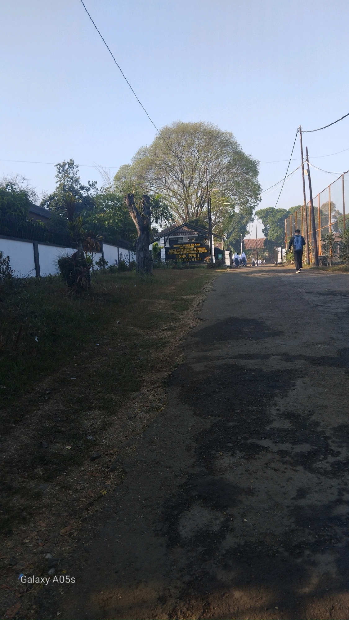 The image shows the entrance to a school in Sumedang, Indonesia. The school's name, "SMK PPB NUSANTARA," is visible on a sign. A young man with a backpack is walking away from the camera towards the school entrance. Several other students are also seen walking towards or standing near the entrance. The school is surrounded by a white wall, and a large tree is prominently featured in the background. The scene appears to be in the morning, given the clear sky and the quality of light. The road leading to the school is unpaved and has patches of dark soil and gravel. The overall environment suggests a rural or semi-rural setting.