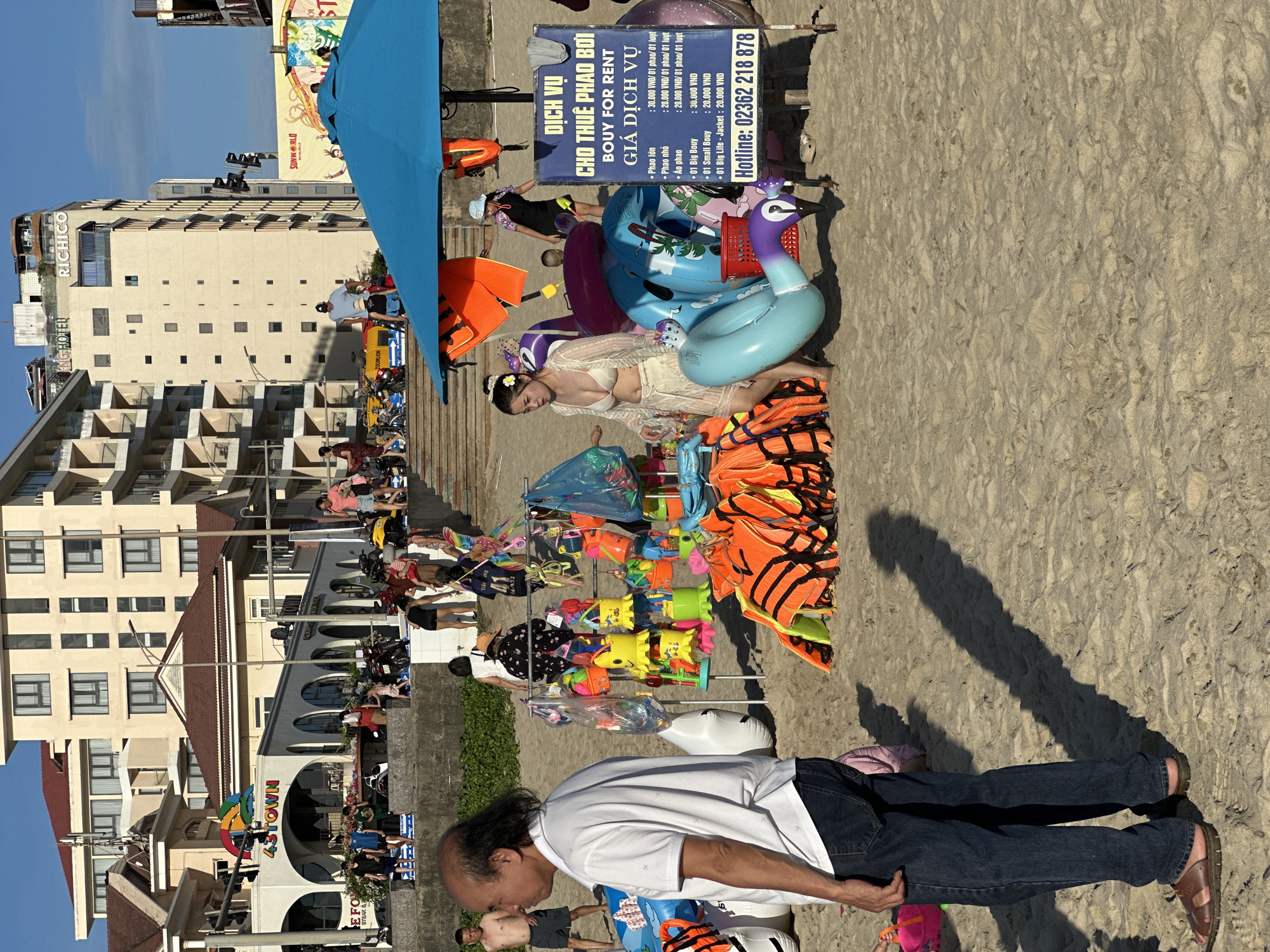 The image depicts a beach scene in Da Nang, Vietnam, likely during daytime given the shadows and the bright sunlight. In the foreground, a man with dark hair and wearing a white polo shirt and blue jeans stands facing left, looking out at the scene. To his right, a rack of colorful beach toys and inflatable rings is displayed, with a stack of orange life vests to its right. A blue umbrella shades some of these items. A woman in a bikini and a sheer cover-up walks past the display. Behind them, a crowd of people are visible on the sand and on a stepped embankment. Further in the background, buildings, including a multi-story hotel with balconies, are visible. Signs in Vietnamese are present, advertising services such as "DICH VU CHO THUE PHAO BOI" (Bouy for Rent Service). The overall atmosphere is that of a busy tourist beach.