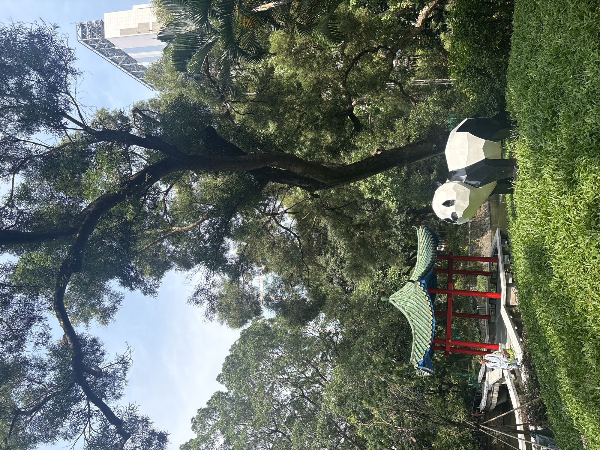 The image captures a scene in Hong Kong, China. A polygonal panda statue stands in front of a building. A traditional Chinese pavilion with red columns and a green tiled roof is nearby, along with a pond. Lush greenery, including trees and bushes, fills the surrounding landscape. The sky is visible through the trees and is blue. A tall building can be seen through the trees.
