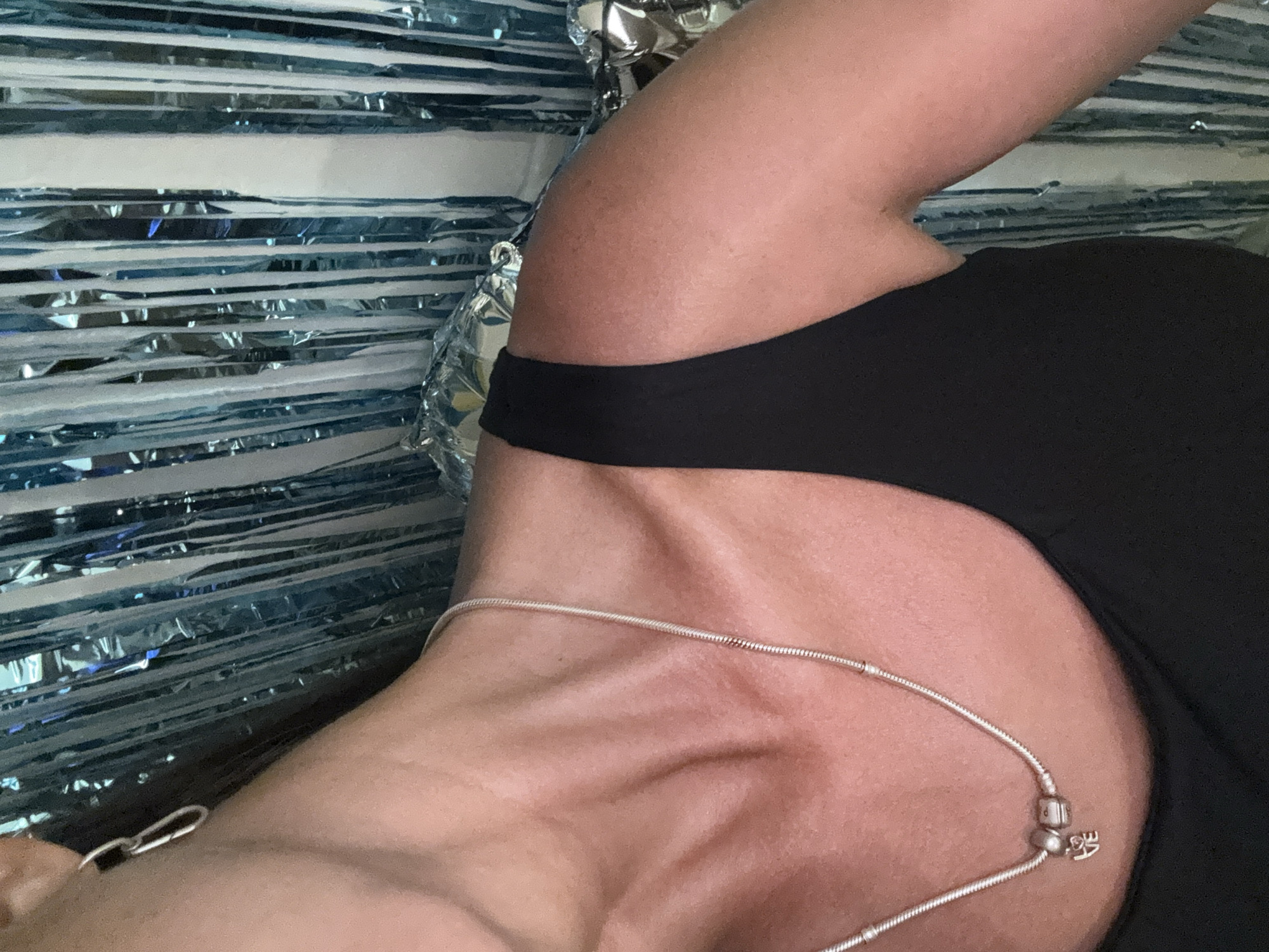 The image captures a close-up, high-angle view of a person, focusing on their neck, collarbone, shoulder, and upper arm. The person is wearing a black top with a broad strap visible over the shoulder. A silver-colored chain or necklace is draped across their collarbone, with a small, spherical bead and a charm visible near the shoulder. The charm appears to have the letters "VE" or "LOVE" etched into it.
The background consists of multiple horizontal strips of shiny, metallic material, alternating between blue and silver or highly reflective silver, suggesting a backdrop of tinsel or foil streamers. This creates a shimmering, patterned effect behind the subject. The lighting seems soft, highlighting the contours of the skin and the reflective nature of the background.
There is no specific activity depicted, but the pose suggests a moment of rest or being photographed. The image conveys a relaxed atmosphere, possibly indoors. The skin tone of the person appears healthy and slightly tanned. Based on the provided context, the location is Kuta Selatan, Indonesia.