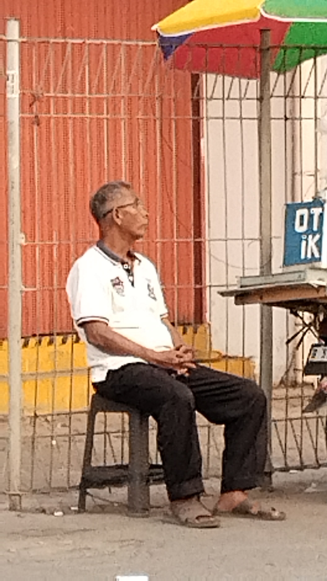An elderly Indonesian man sits on a dark stool on a sidewalk in West Jakarta, Indonesia. He is wearing glasses, a white polo shirt with a dark collar, and dark pants. His hands are clasped in his lap. Behind him is a red metal fence. A multi-colored umbrella is visible above and to the right of the man. A table with a blue sign featuring white text is to his right. The ground appears to be paved.