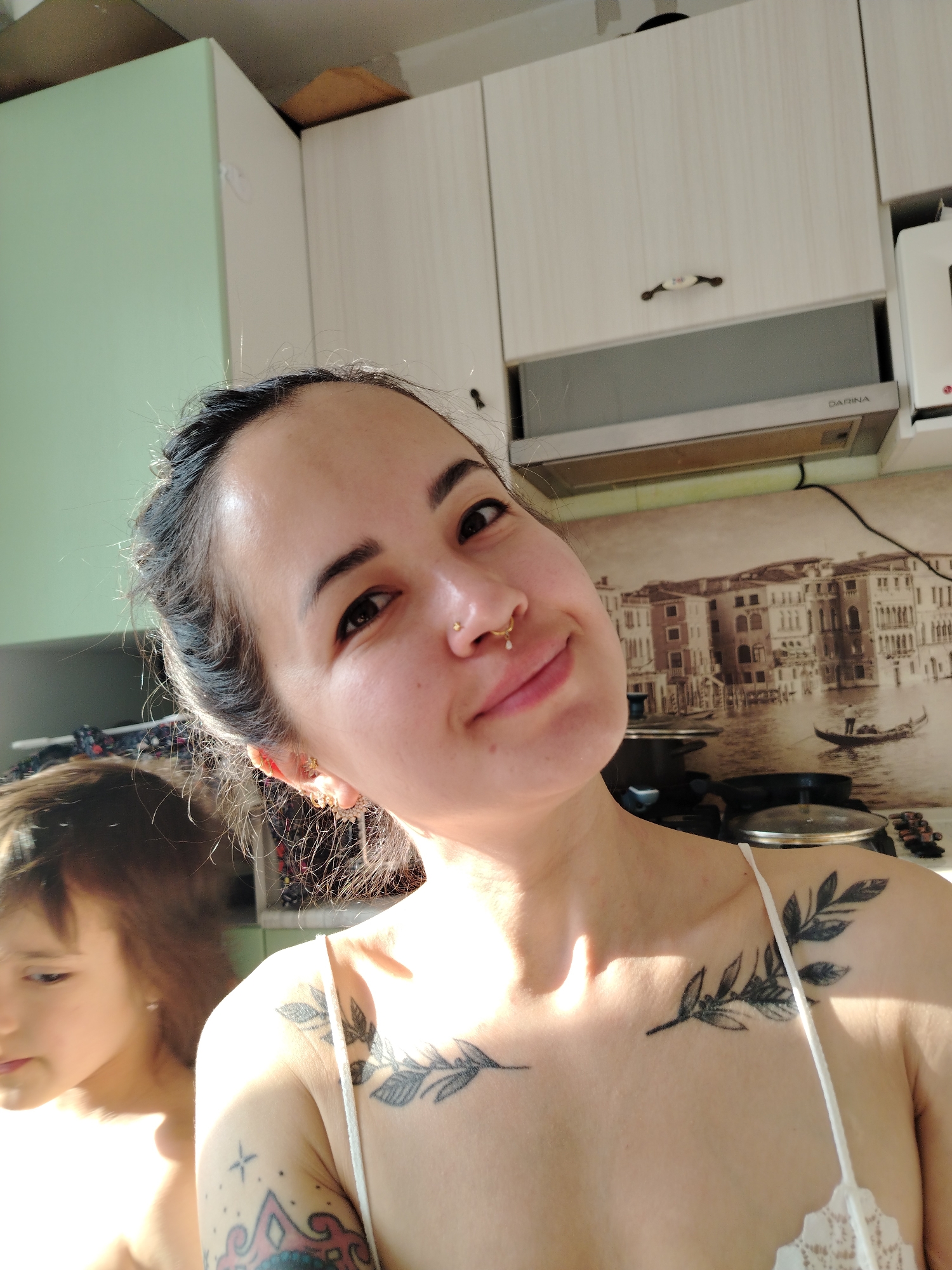 A woman is taking a selfie in a kitchen. She has dark hair pulled back, pierced nose, earrings, and tattoos on her chest and arm. Reflected in a mirror next to her is a child with brown hair. In the background are kitchen cabinets, a range hood labeled "DARINA", and a wall covering featuring a Venice canal scene. There are pots and pans visible on the counter. The location is Noviy, Russia.