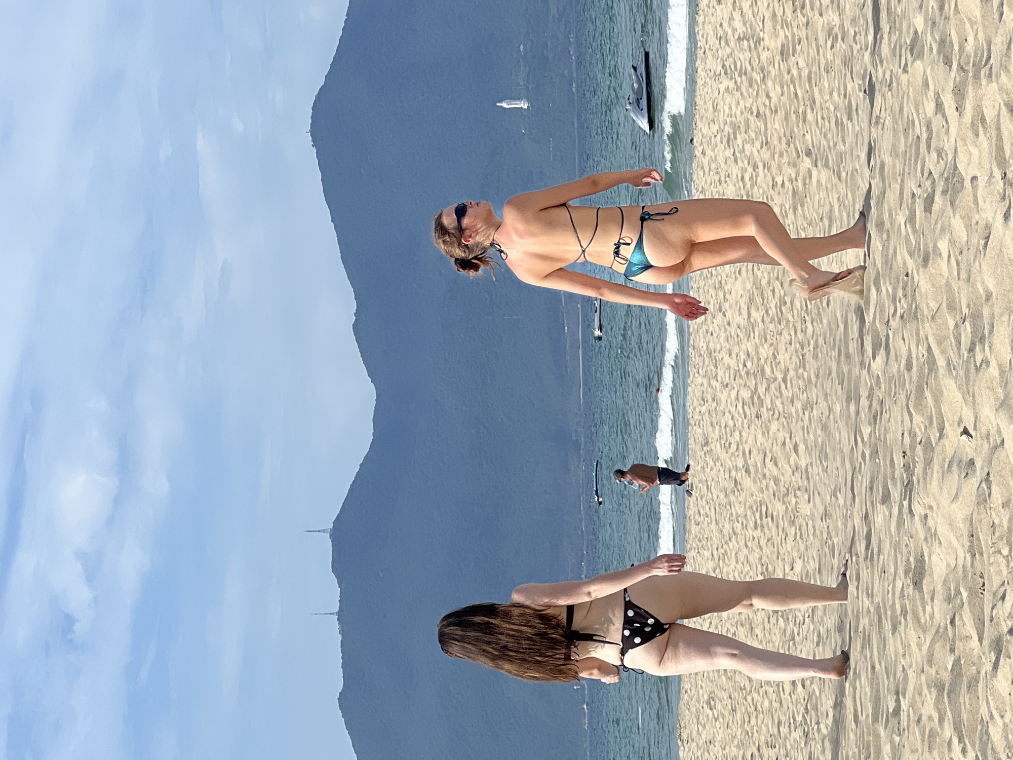 Two women are walking on a sandy beach in Đà Nẵng, Vietnam. In the background, the sea is visible, with several boats and a jet ski. Behind the sea, a mountainous landscape is present with two prominent radio towers on one of the peaks. A large white statue is visible on a hill on the right side of the image, overlooking the sea. The sky is blue with scattered clouds.