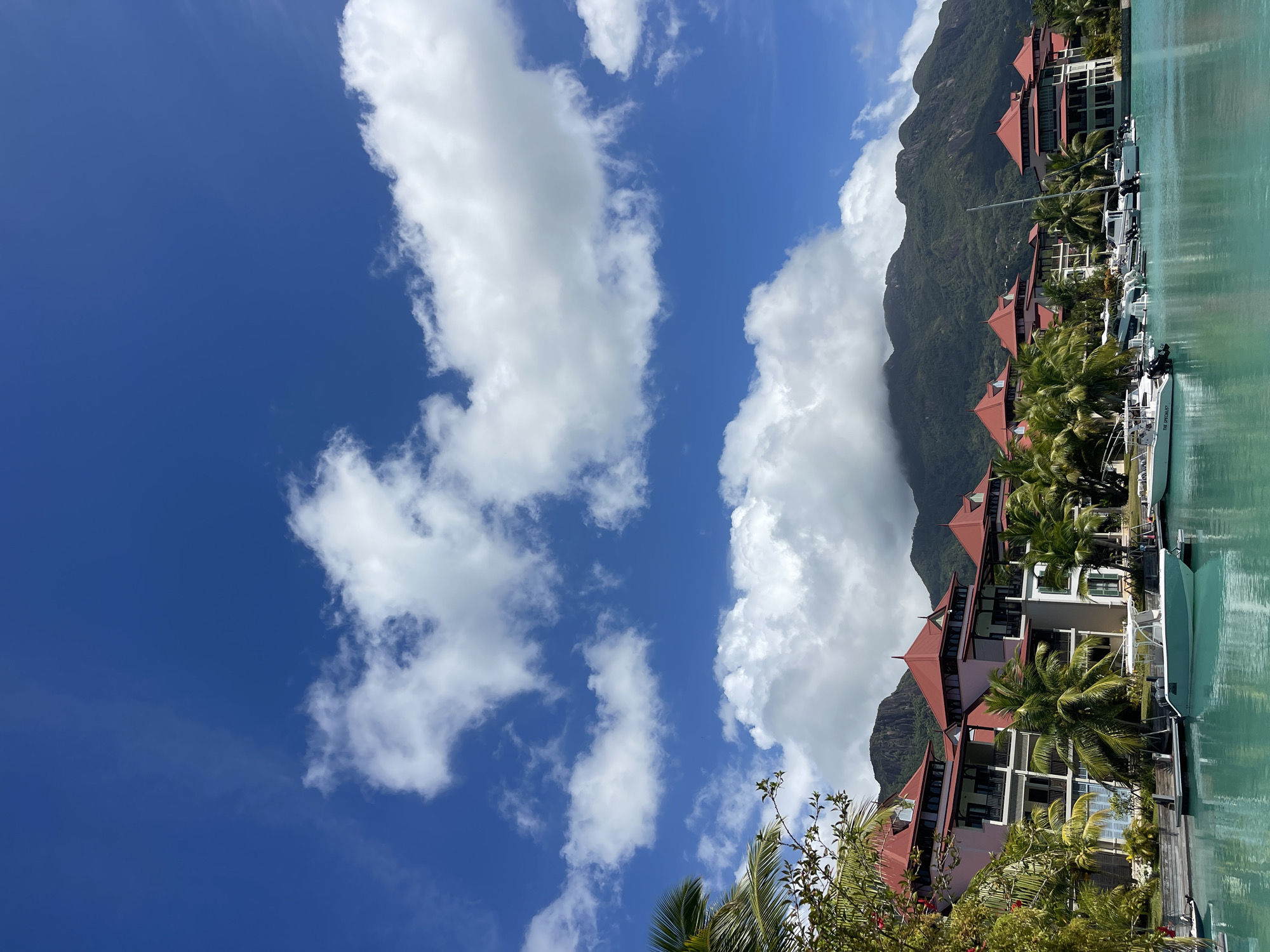 A wide-angle, vertical shot captures a tropical resort scene in Victoria, Seychelles, under a bright blue sky dotted with fluffy white clouds. In the foreground, turquoise water laps against a wooden dock where several white motorboats are moored. Behind the boats, a multi-story resort building with distinctive red-tiled roofs and pink-accented walls stretches across the frame. Palm trees and lush green foliage surround the building, adding to the tropical ambiance. In the background, verdant, cloud-shrouded mountains rise dramatically, forming a picturesque backdrop to the resort. The scene is tranquil and idyllic, evoking a sense of vacation and relaxation.