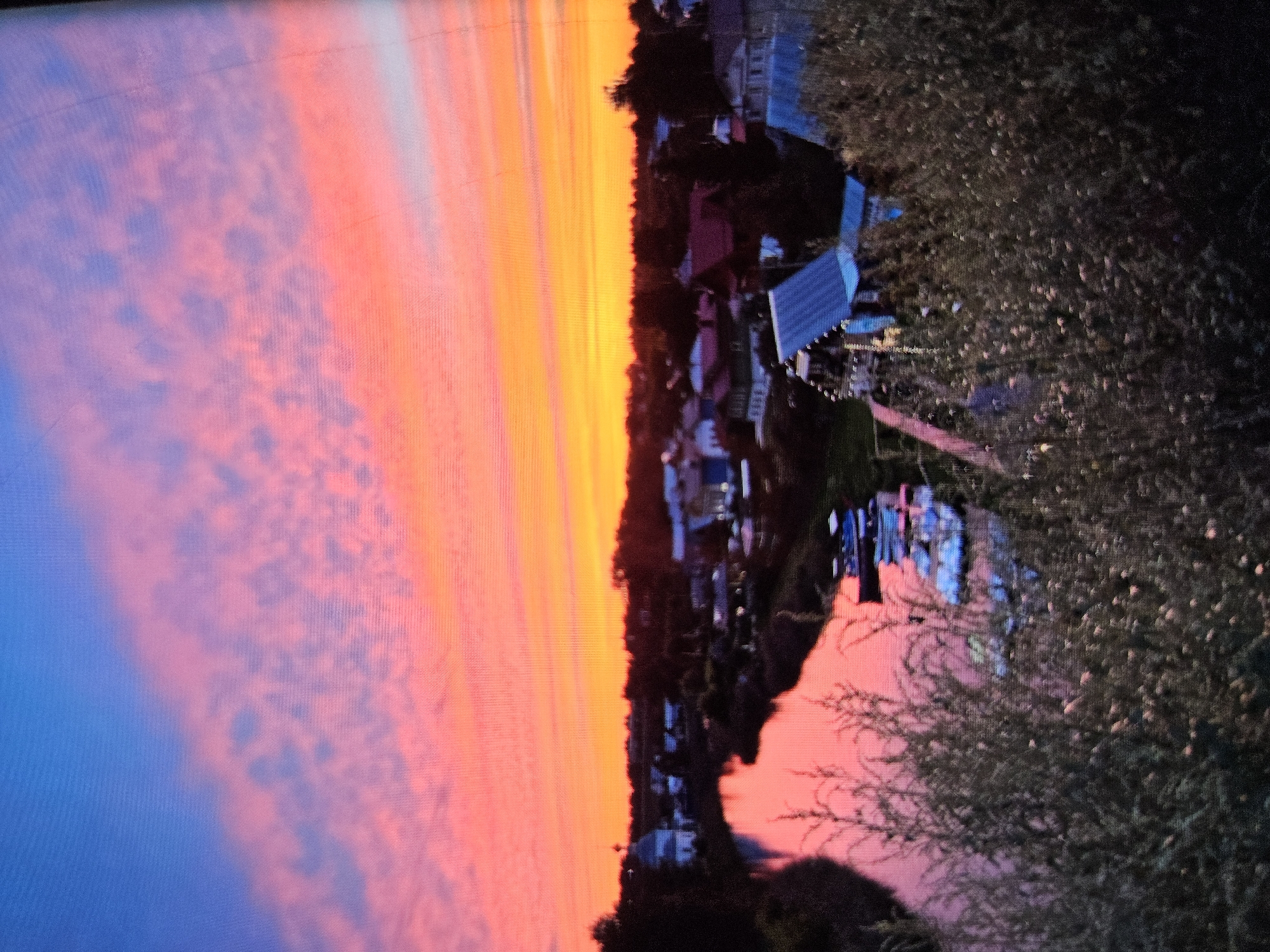 The image depicts a sunset over a rural settlement, possibly in the city of Vladimir, Russia, given the presence of traditional Russian architecture, including a prominent church with a dome. The sky is ablaze with vibrant colors of pink, orange, and yellow, transitioning to blue at the top, suggesting a time around dusk. A river flows through the scene, reflecting the colorful sky. The riverbank is lined with trees and a cluster of houses, some with distinctive red roofs, characteristic of Russian villages. In the foreground, there is dense foliage and several boats moored along the river. The overall impression is one of a peaceful, picturesque rural landscape bathed in the warm glow of the setting sun.