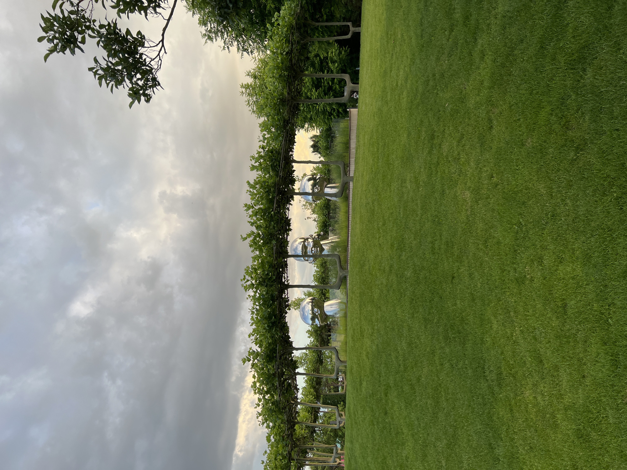 The image shows a park-like outdoor scene with a large expanse of well-maintained green lawn in the foreground. In the middle ground, there is a row of sculpted trees with their branches trained horizontally to form a canopy. Beneath this canopy, several large, polished chrome sculptures of human heads are visible. The heads are smooth and reflective, capturing the light from the sky. The background features more trees and vegetation, and the sky is overcast with dramatic clouds, suggesting either early morning or late afternoon. The scene appears to be a modern art installation within a landscaped garden. The location in Krasnodar, Russia, is identified as the Park of Galitsky (also known as Krasnodar Park).