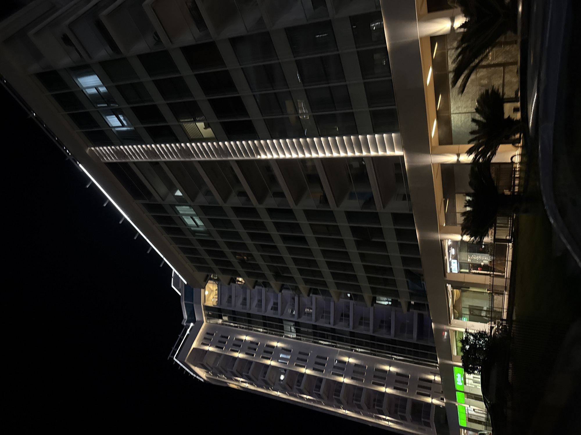 A night-time, low-angle, full shot depicts two modern, multi-story buildings situated in Budva, Montenegro. The building on the left is lighter in color and features numerous balconies with glass railings, illuminated by vertical rows of warm-toned lights. The building on the right is darker and appears to have a facade composed primarily of glass windows, with a vertical strip of integrated, segmented lighting running down its center. Both buildings are topped with subtle exterior lighting. The lower levels of the buildings feature storefronts with illuminated signage, including one with a bright green sign. Palm trees are visible in the foreground, along with a dimly lit fence and the silhouette of a parked vehicle. The overall scene suggests a commercial or residential complex in an urban setting at night.