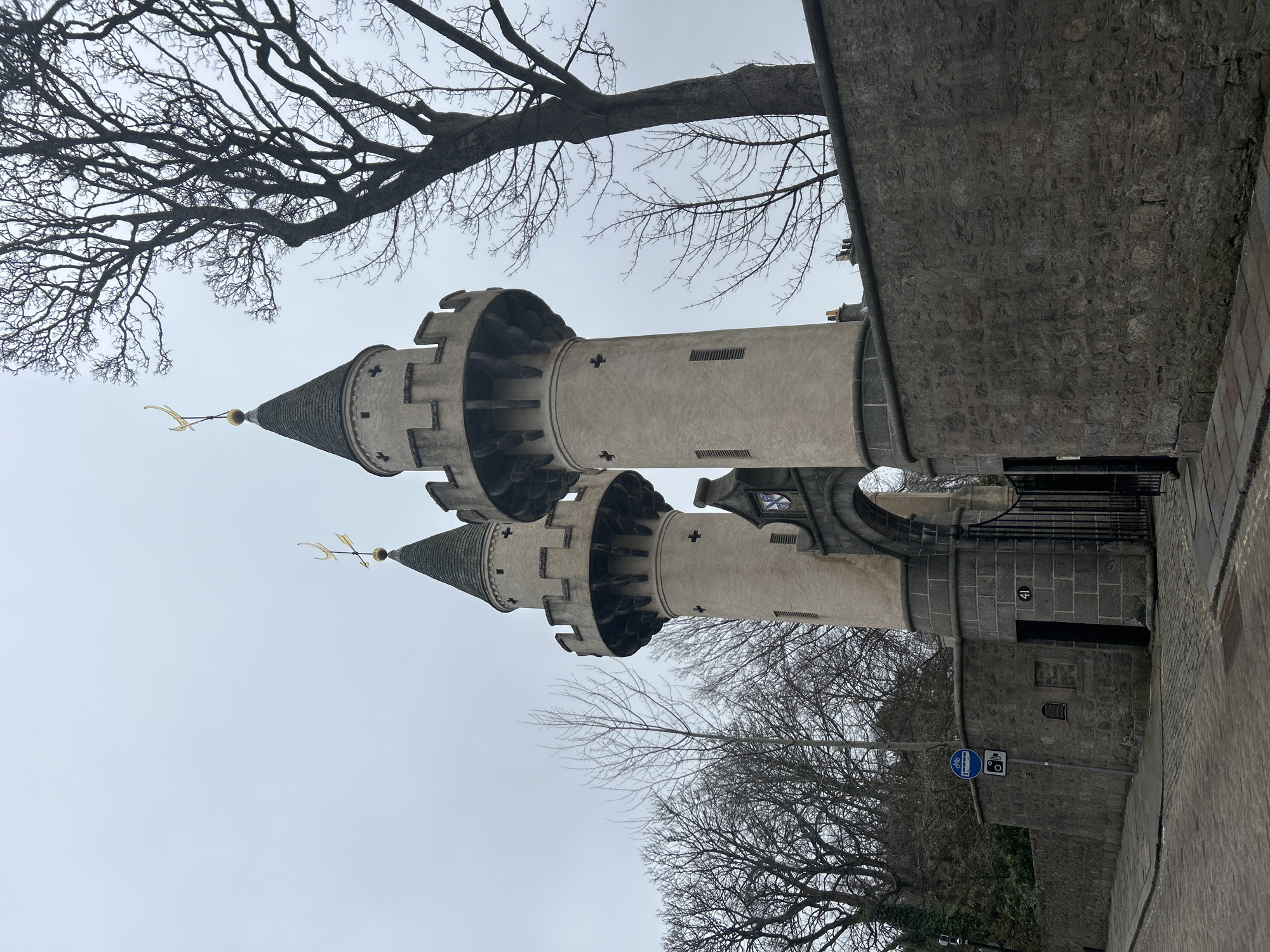 The image shows the towers of St. Machar's Cathedral in Aberdeen, Scotland, United Kingdom. The two prominent towers are made of light-colored stone with decorative features and conical roofs topped with weather vanes. Bare tree branches frame the towers against a cloudy sky. Part of a stone wall extends from one of the towers and runs alongside a paved road. A signpost with a blue circular sign displaying bicycle symbol and a rectangular sign with an image of a camera above a coin slot is also visible near the base of the tower.