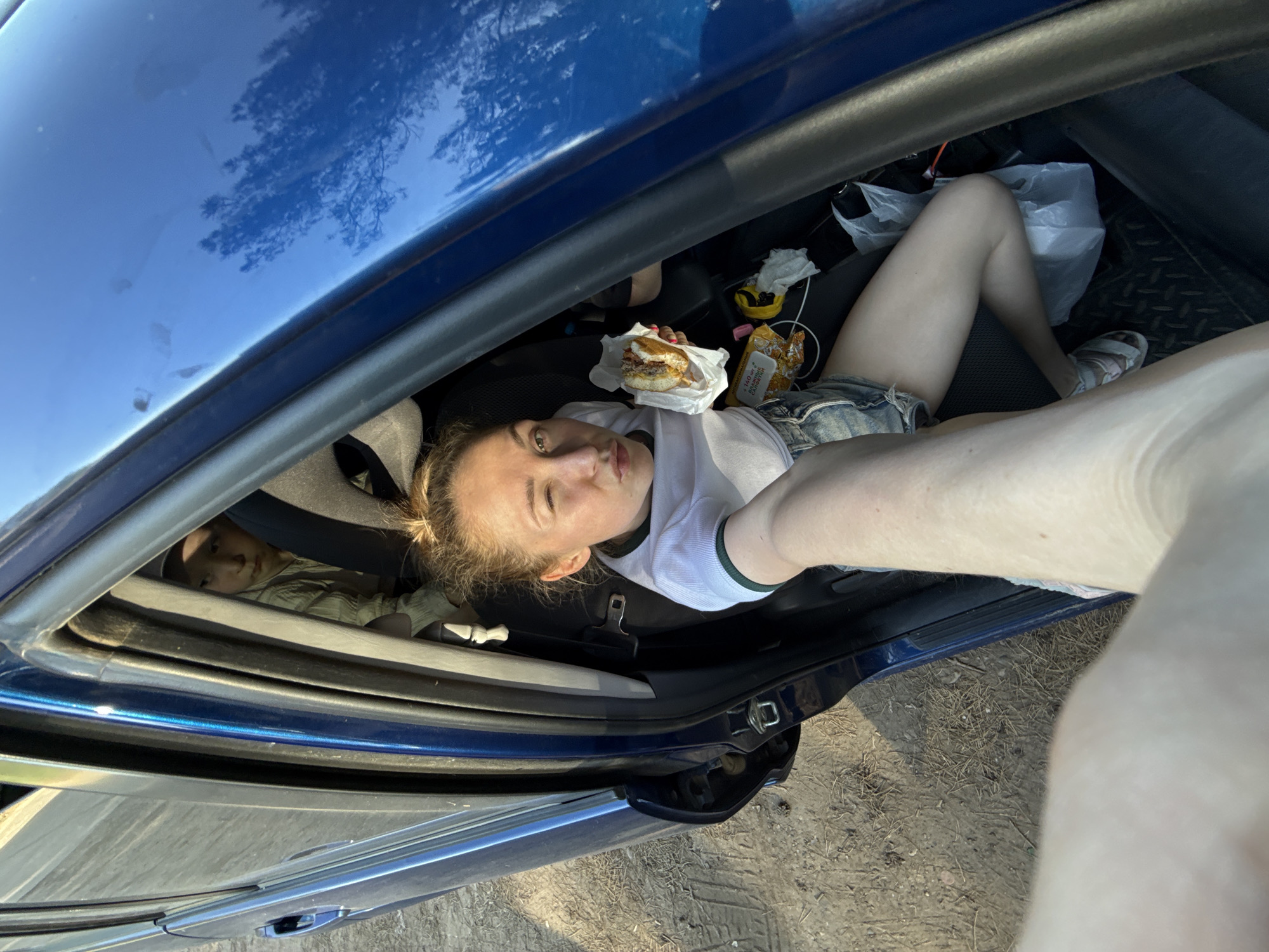 The image appears to be taken inside a blue car, viewed from an unusual angle. A young woman is lying in the back seat. She is holding a partially unwrapped burger in her left hand. There's fast food packaging in the vicinity. Another young child lies in the backseat facing the woman. The car's surroundings show possible dirt, branches or weeds. The image doesn't show sufficient details to determine if the image was taken in the city of Chaikovsky, Russia.