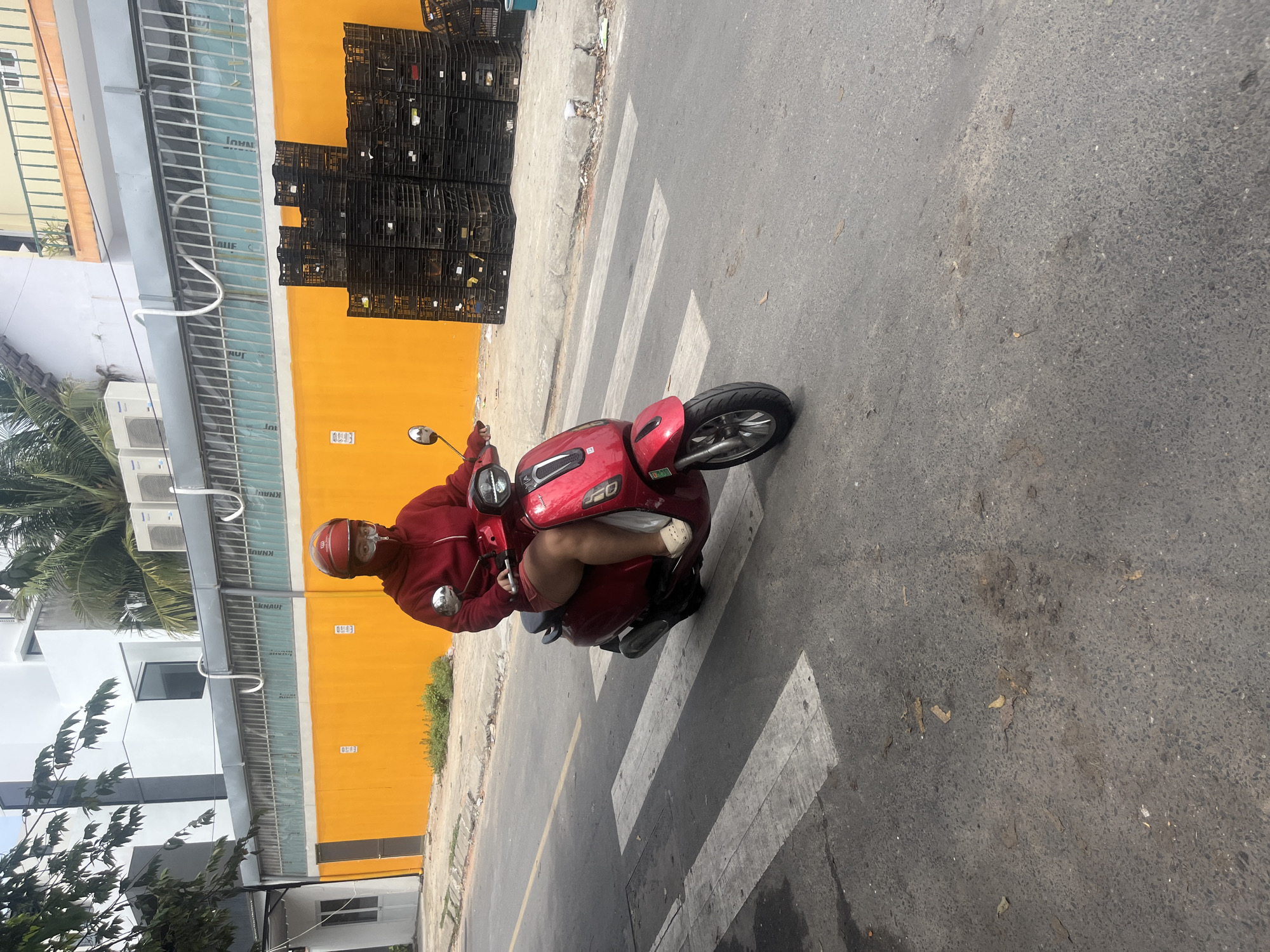 In an urban setting in Da Nang, Vietnam, a person on a red scooter is depicted riding on a street. The rider is wearing a red hooded jacket, a red face mask, and a helmet, obscuring most of their face. They are also wearing light-colored shoes and shorts. The scooter is a modern model, with a prominent front light and silver handlebars. The street features a pedestrian crossing marked with white stripes. In the background, a bright orange wall forms the facade of a building, with several stacks of black plastic crates placed along the sidewalk in front of it. Above the orange wall, a building with white and gray elements and palm trees are visible. The sky is overcast, suggesting a cloudy day. The overall scene captures a slice of daily life on a city street, with the rider navigating the urban environment.
