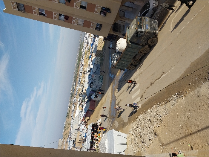 A truck carrying what appears to be water or supplies drives along a road bordering a densely populated area of tents and makeshift shelters. People walk along the road near the truck. Tall buildings frame the scene. The sky is hazy with sunlight. The ground around the road appears arid and partially developed with gravel and dirt.