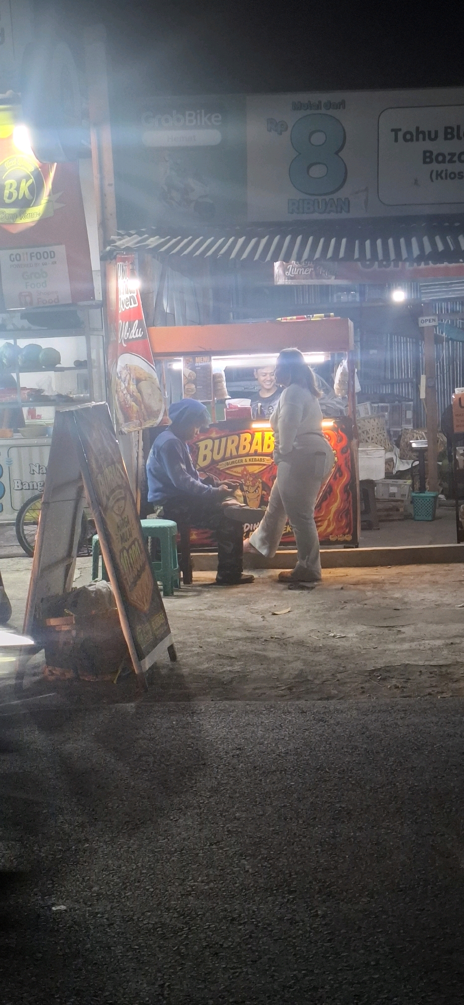 The image shows an outdoor scene at night in Pabelan, Indonesia, likely near a food stall. Two individuals are present. One, wearing a blue head covering and a blue jacket, sits on a green stool, looking down at a mobile phone. The other, wearing light-colored pants and a light-colored top, stands, with their right leg extended forward. They are positioned in front of a food stall with a banner that reads "BURBAB" and "BURGER & KEBS." The stall has a menu displayed and appears to be illuminated by overhead lighting. In the background, there are signs for "GrabBike" and "Tahu Bl..."; the latter appears to advertise a price of 8 Ribuan. The ground is paved with asphalt and concrete, with scattered debris. A wooden sign stands to the left of the individuals, partially obscured. The overall lighting is dim, with light sources creating lens flares and affecting visibility.