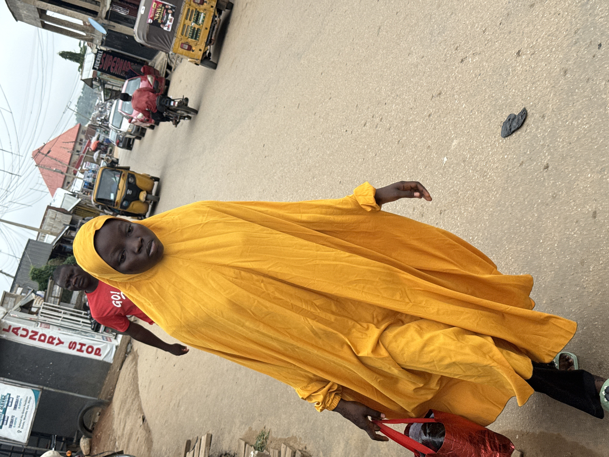 A woman in a bright yellow hijab and dress walks on a paved street in Abuja, Nigeria, carrying a red bag. Her head is bowed, and her expression is somber. Behind her, a man in a red shirt with the word "GO!" printed on it walks with a determined stride. The street is lined with buildings, some appearing to be shops, with a "LAUNDRY SHOP" sign visible on one. Several vehicles are present, including a yellow three-wheeled auto-rickshaw, a motorcycle with a rider, and a few cars. The sky is overcast, suggesting a cloudy day. The overall scene depicts a busy street in an urban environment.