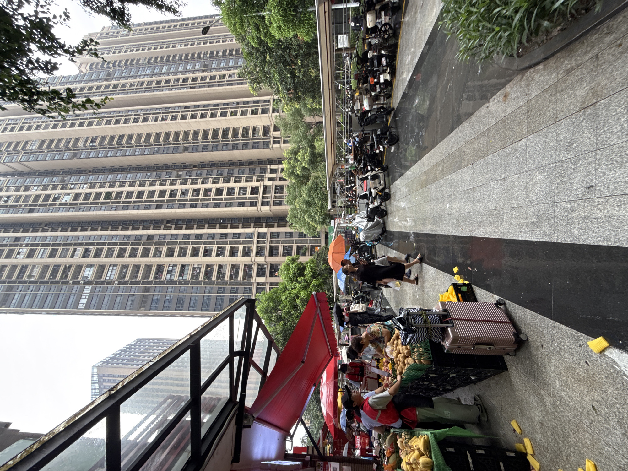 In Haizhu District, China, a street market scene unfolds with tall residential buildings in the background. A covered stall features a vibrant red awning and displays various fruits and vegetables, including bananas and potatoes, in crates. Several individuals are present, including vendors attending to customers and shoppers browsing the produce. Some people are holding umbrellas, suggesting it may be raining, as indicated by the wet pavement. Adjacent to the market, a designated parking area is filled with numerous motorcycles and scooters. A pink suitcase rests on the ground near the produce stall. The scene depicts a bustling urban environment with commercial activity and typical city infrastructure.