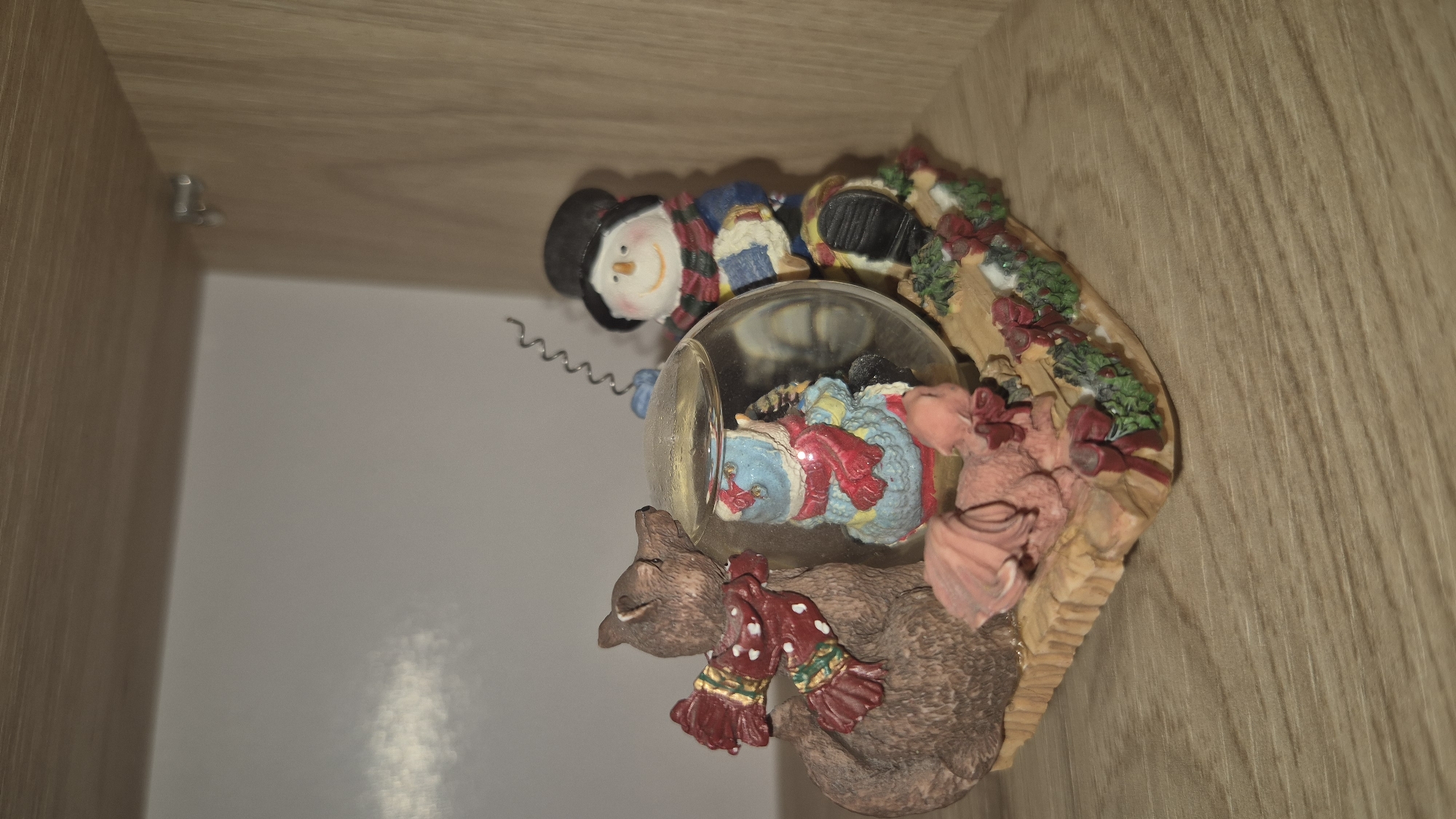A resin figurine depicts a winter scene with a brown bear, a snowman, and a small pink dragon. The bear, wearing a red scarf with white polka dots and green trim, is to the left of a glass snow globe. Inside the globe, a blue and white figure, possibly another snowman or penguin, is visible. To the right of the globe stands a snowman wearing a black top hat, a blue coat with a checkered scarf, and holding a coiled wire above his head. A small pink dragon lies on the wooden base in front of the snow globe. The base is decorated with greenery and red bows, resembling a wreath. The figurine is placed on a wooden shelf within a light-colored enclosure, suggesting it is part of a display. The environment does not provide any indicators of location within the city of Butovo, Bulgaria.