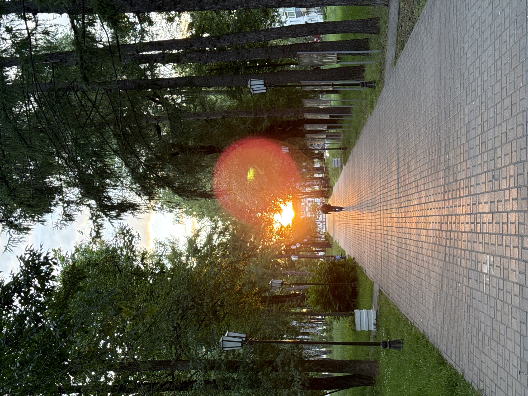The image depicts a park pathway in Chaikovsky, Russia, during what appears to be sunset. A person is walking away from the camera along the paved path, which is bathed in the warm, golden light of the setting sun. The path is lined with trees and lampposts, creating a serene and inviting atmosphere. The sun is low in the sky, casting long shadows and creating a lens flare effect. The sky is partly cloudy, with hints of orange and yellow from the sunset. The presence of lampposts suggests this is a public park or a pedestrian area designed for evening strolls. The overall mood is peaceful and tranquil, evoking a sense of calm and natural beauty.