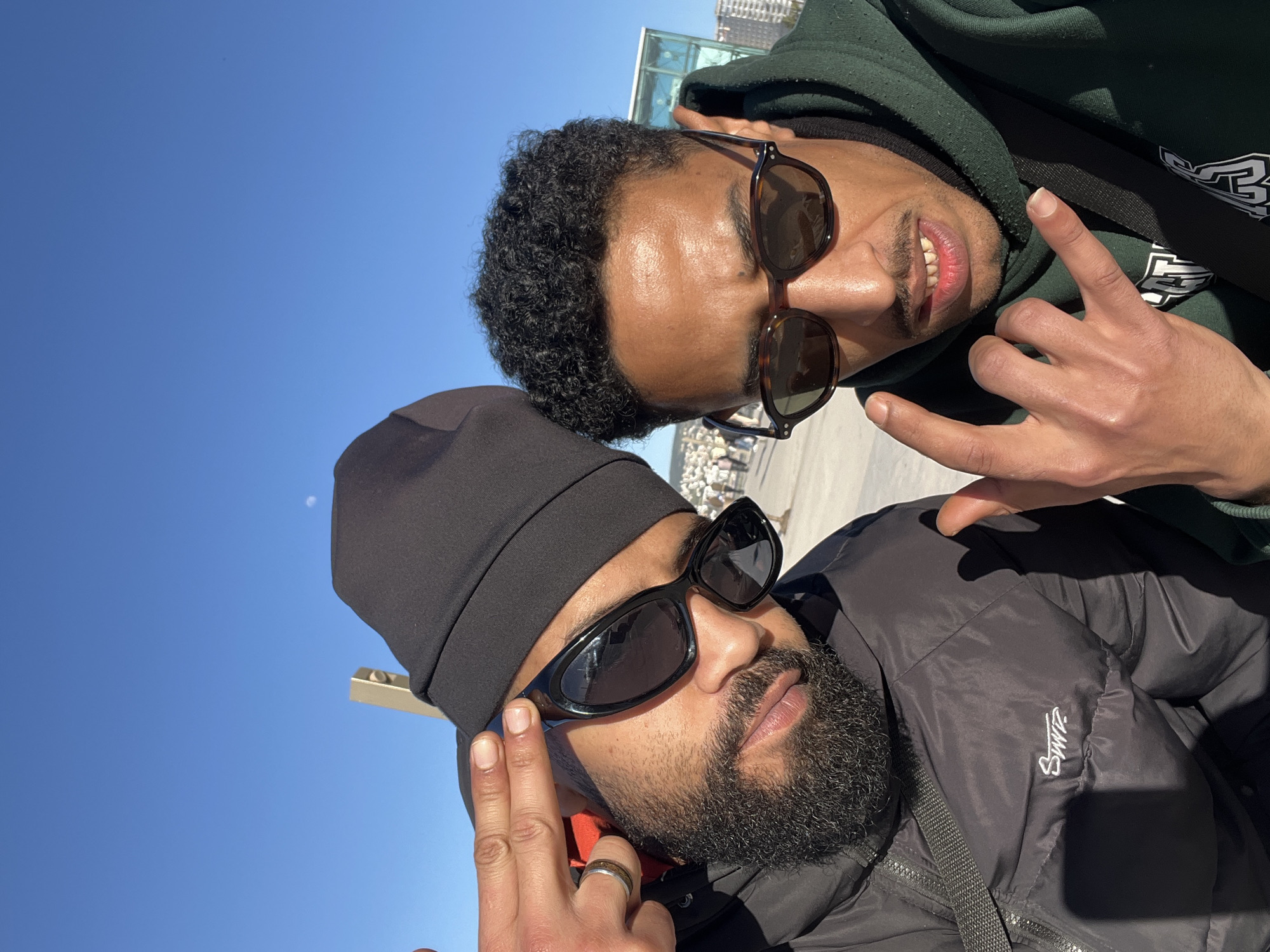 Two men pose for a selfie against a bright blue sky, likely in Tanger, Morocco. The man in the foreground wears a black beanie, dark sunglasses, and a black puffer jacket with a white logo on the sleeve. He has a full beard and is making a hand gesture with his fingers. The man behind him wears a dark green hoodie with a white design, and tortoiseshell sunglasses. He is also making a similar hand gesture. In the blurred background, there is a metal railing, some buildings, and a crowd of people.