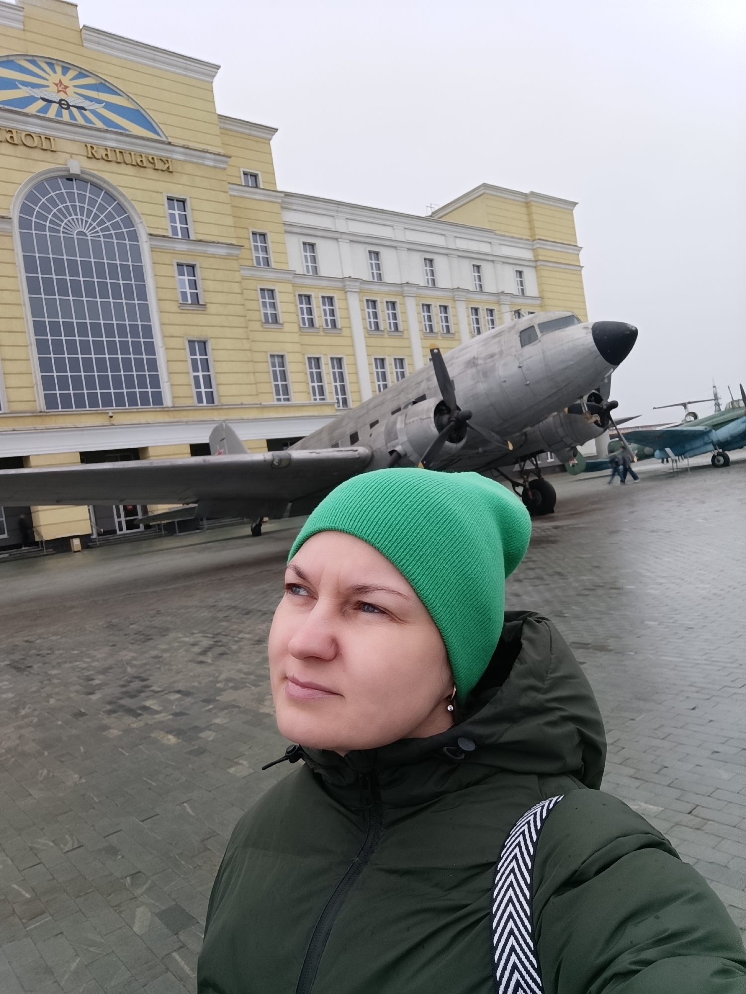 A woman wearing a bright green beanie and a dark green puffer jacket is the central subject of the image, appearing to take a selfie or be photographed. She has a neutral, contemplative expression, looking towards her left.
Behind her stands a large, silver-grey twin-engine propeller aircraft, resembling an Li-2 (Soviet license-built Douglas DC-3), with its nose and propellers prominently visible. Further in the background, another, smaller blue-green aircraft, possibly a biplane, is also on display, with a few people visible near it. A faint red star emblem can be seen on the tail of one of the aircraft.
The backdrop features a substantial yellow building with white architectural details, including cornices and window frames. Its left side boasts a large arched window with a grid pattern. Above this window, a distinctive blue and yellow emblem with a red star and stylized wings is mounted. Cyrillic text is visible on the building's facade, which reads "МУЗЕЙ УГМК" (UMMC Museum).
The ground is paved with dark grey and black rectangular tiles, appearing slightly damp. The sky is overcast and grey, suggesting a cool, cloudy day. The scene depicts an outdoor exhibition, likely an aviation museum, specifically the UMMC Museum of Military Equipment in Verkhnyaya Pyshma, Russia.