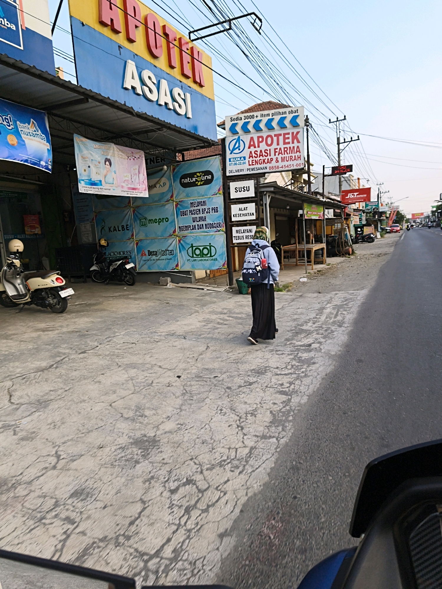 A person wearing a hijab and a long black skirt walks on a cracked concrete sidewalk next to a road in Desa Ngesrep, Indonesia. The person is carrying a backpack and walking away from the camera. In the background, there are several shops and a pharmacy with banners and signs. One sign reads "APOTEK ASASI FARMA LENGKAP & MURAH" (Asasi Pharmacy, Complete & Cheap). There are also motorcycles parked on the sidewalk. The sky is clear and the sun is shining. The road is paved and there are cars and motorcycles on it.