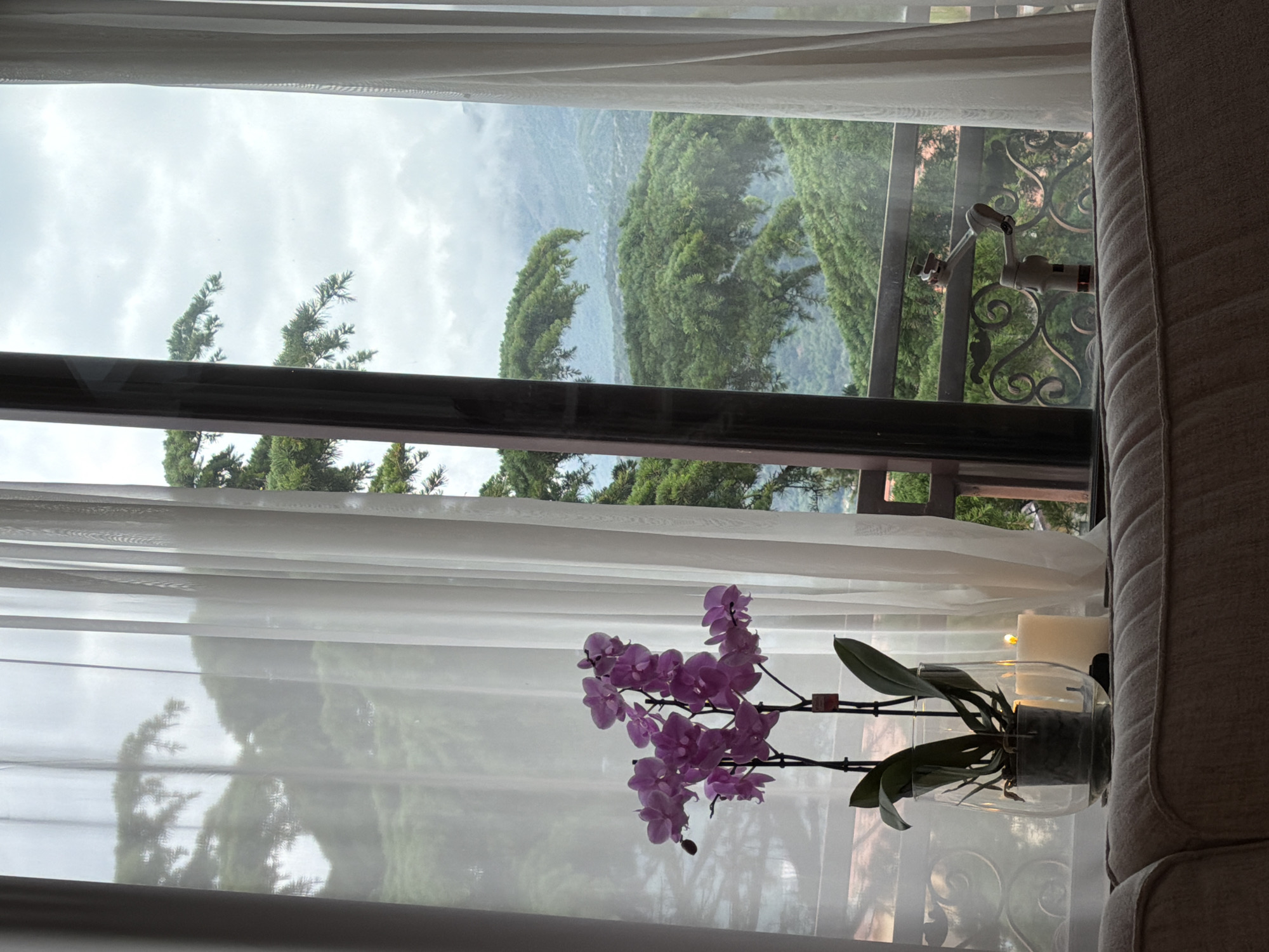 A view from inside a room, partially obscured by a white curtain, reveals a verdant outdoor landscape. A window frame outlines a view of rolling hills densely covered with green vegetation. The sky is overcast, casting a diffuse light over the scene. In the foreground, an orchid plant with light purple flowers sits in a glass vase on a window sill. Next to the plant are two white candles of varying sizes. To the right, the side of a light-colored sofa is partially visible. A balcony railing is visible outside of the window. Given the mountainous, coastal setting, it is possible that the location of this image is in Budva, Montenegro.