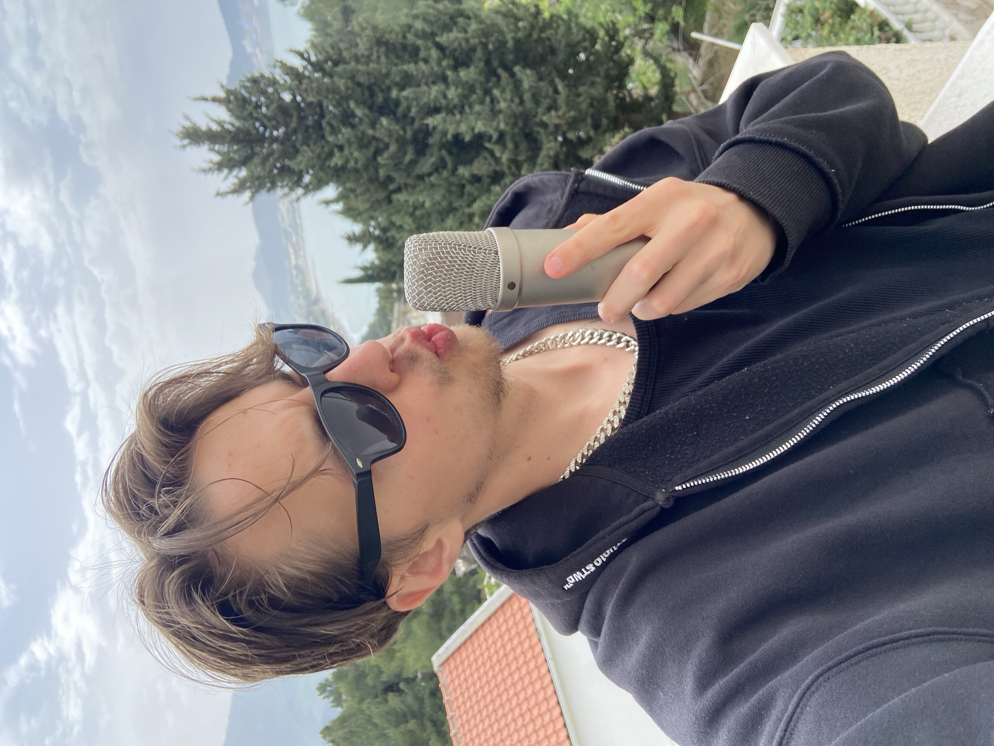 A young man with light brown hair, wearing sunglasses, a black zip-up hoodie, and a silver chain, holds a microphone to his lips. The background shows a tree, water, and mountains, suggesting an outdoor location in Šušanj, Montenegro. The image is taken at an angle. A roof with red tiles is partly visible in the lower part of the frame.