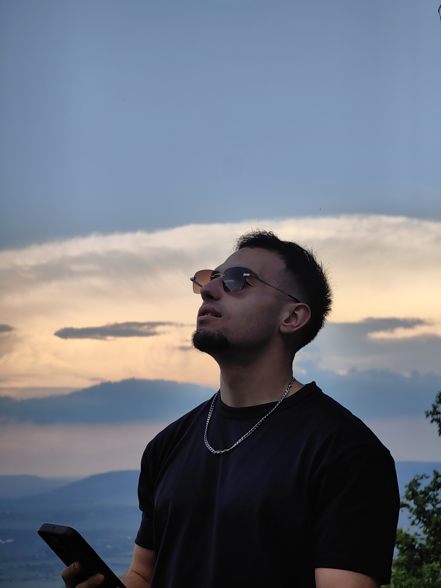 A medium shot depicts a man with tan skin wearing a black T-shirt and a silver chain, holding a black smartphone in his left hand. He is also wearing sunglasses with a brown tint. He has dark hair and a beard, and is looking upwards towards the sky. The background shows a landscape with hills or mountains partially obscured by clouds. The sky is a mix of blues and oranges, indicating either a sunrise or sunset. Although it is not explicitly identifiable from the image, it is described as being in the city of Shumen, Bulgaria.