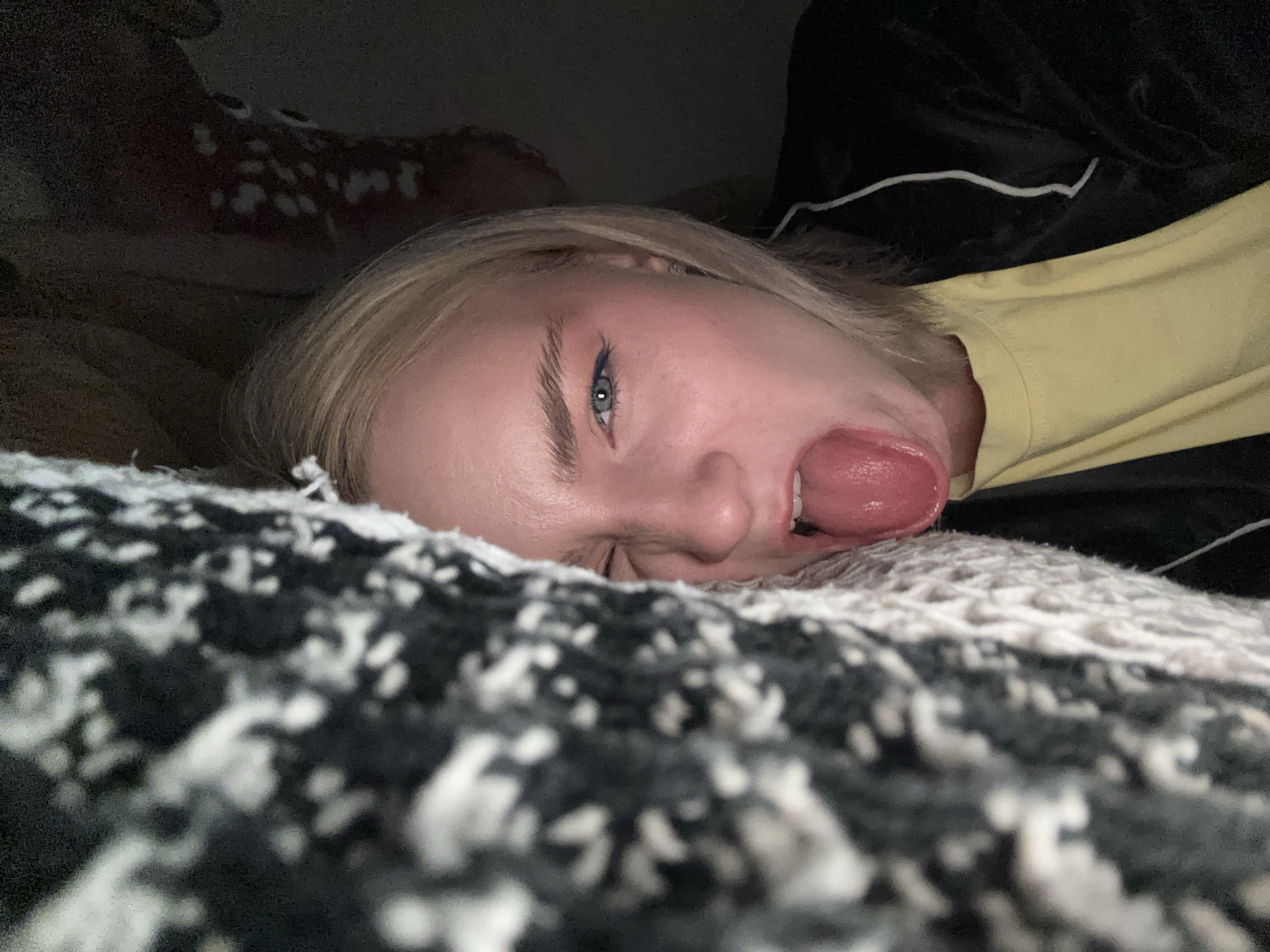 A close-up, low-angle, indoor shot shows a person's face with their tongue sticking out, lying on a patterned blanket. The person has blonde hair and is wearing a yellow shirt and a black jacket with white stripes. The background is dark and out of focus, with a hint of a deer-patterned object. The scene is likely a bedroom or living area. There is no indication of the city of Lednevo, Russia.