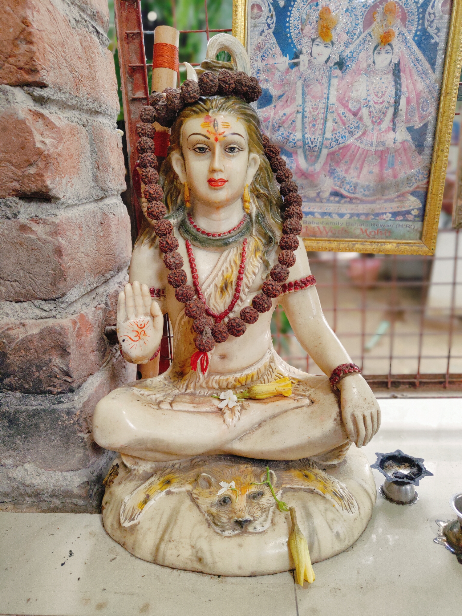 The image shows a statue of Shiva, a Hindu deity, seated in a cross-legged position on a platform depicting a lion. The statue is made of a light-colored material and is adorned with a necklace of rudraksha beads and a red beaded bracelet. Shiva's right hand is raised with the palm facing outward, and the Om symbol is painted on it. A small yellow flower and a green leaf are placed on the statue's lap. Behind the statue, a framed picture of Radha and Krishna is visible. To the left of the statue is a brick wall, and to the right, there are small oil lamps. The scene appears to be an outdoor shrine, possibly located in Gurgaon, India, given the context of the photograph. The floor is tiled and speckled with dirt.