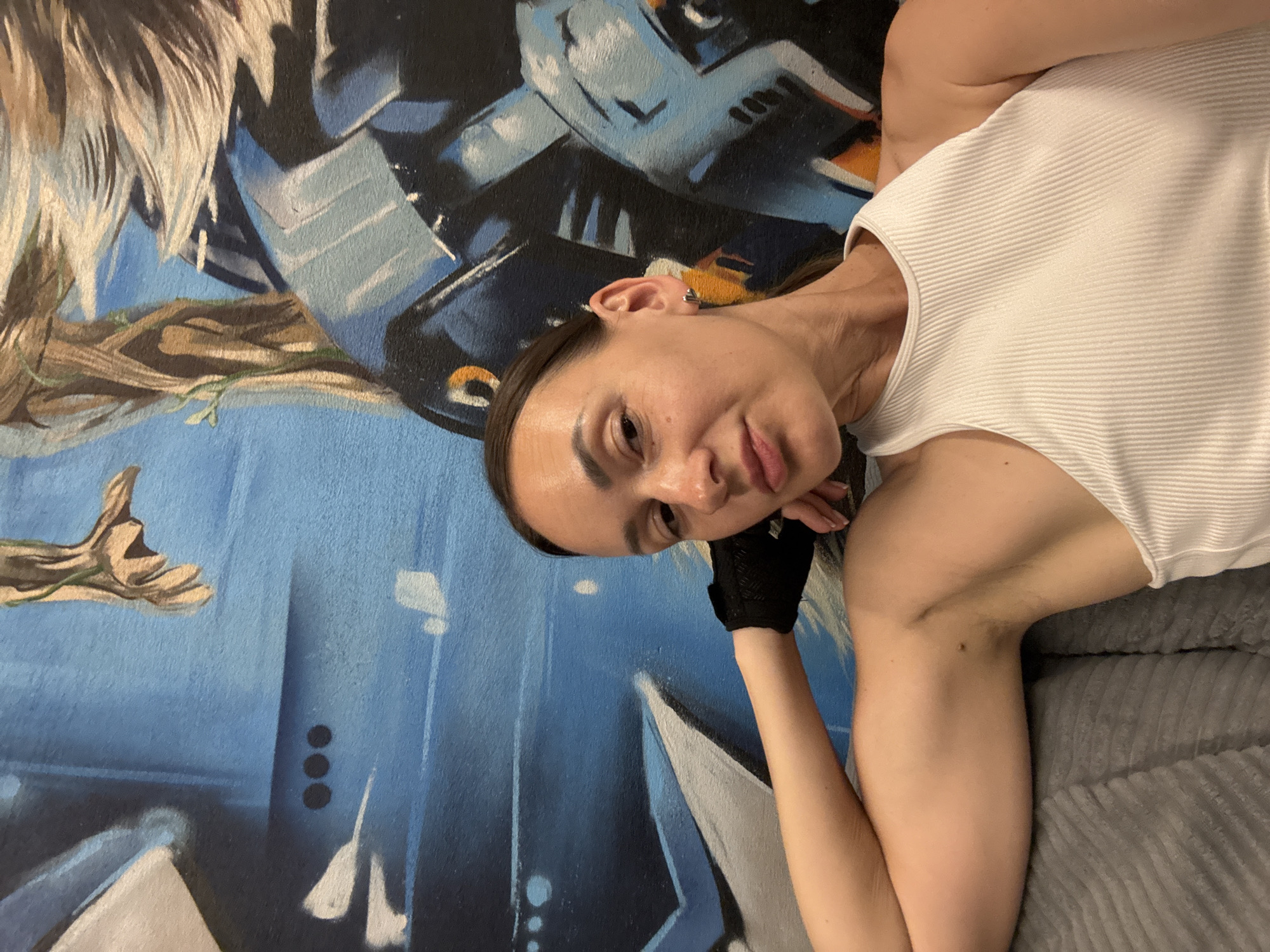 A portrait of a woman lying down with her head resting on her hand, against a blue mural with abstract shapes. The woman is wearing a white ribbed tank top and a black glove on her right hand. Her hair is dark and pulled back. She has an earring in her left ear. The mural behind her is predominantly blue with black and white abstract elements. The image appears to be taken indoors, possibly in a residential setting, given the presence of a textured grey blanket or couch beneath her. The lighting suggests it is daytime. The woman's expression is calm and relaxed. No visible text is present in the image. The context is Sochi, Russia.