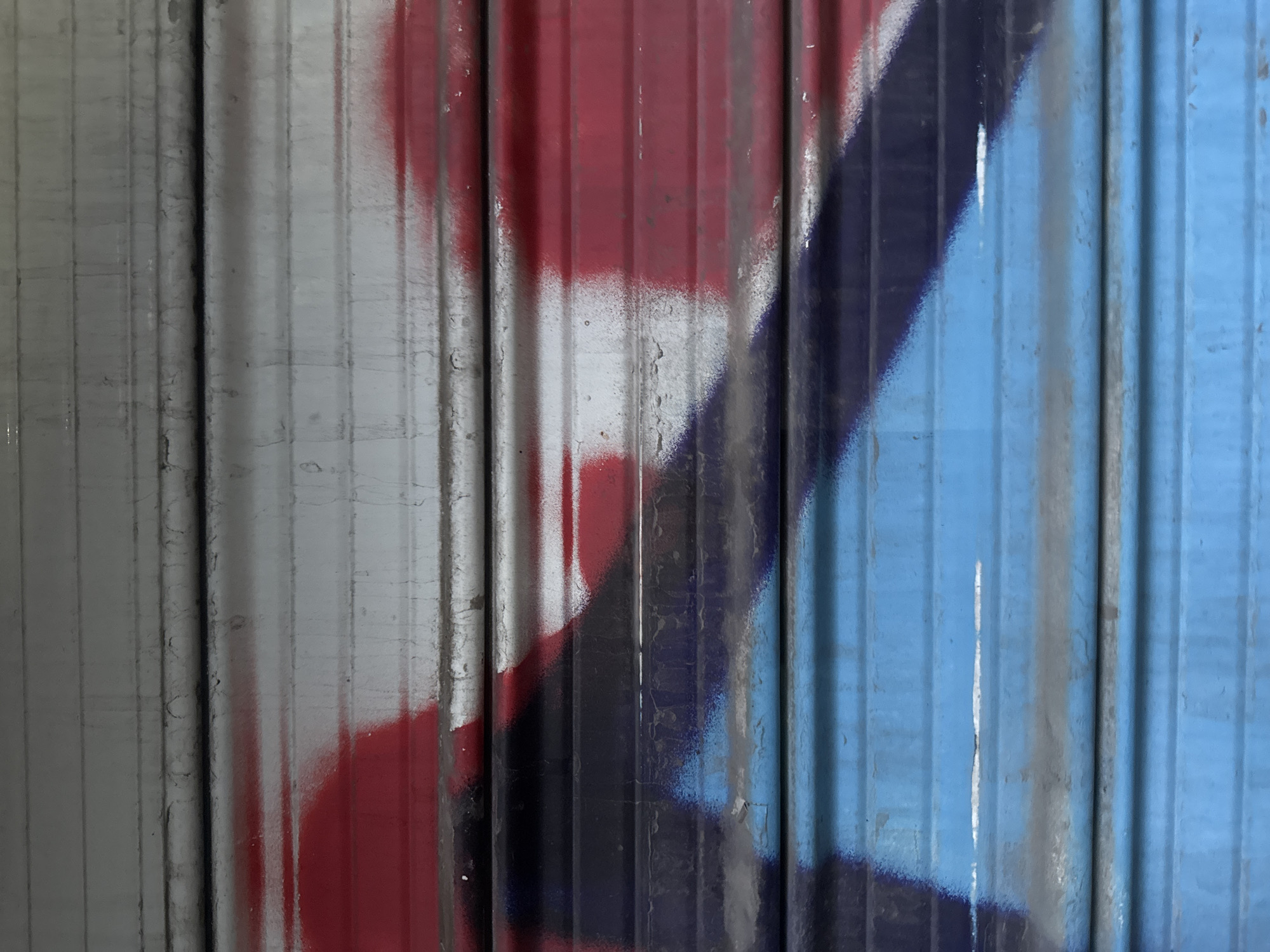 The image shows a section of a corrugated metal wall or door, heavily marked with spray paint in shades of gray, red, blue, and black. The paint is applied haphazardly, creating an abstract design with blurred edges and inconsistent coverage. The metal surface itself exhibits signs of age and wear, including streaks and minor surface imperfections. The corrugated pattern runs vertically, emphasizing the linear nature of the image. No people are visible. The location is unknown, and there is no information to suggest this image was taken in Sofia, Bulgaria.