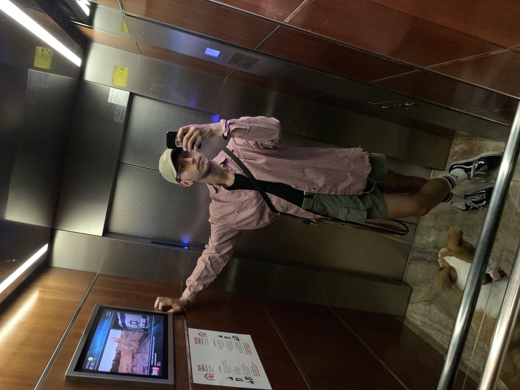 The media file captures a selfie taken by a person in an elevator, which is evident from the highly reflective walls and the person holding a phone pointed towards a mirror. The image is taken at an angle, with the person appearing to lean against the elevator wall.
The main subject is a young male wearing a light-colored baseball cap, yellow-tinted sunglasses, a black t-shirt, a red and white striped long-sleeved button-down shirt, green shorts, white socks with black stripes, and black sneakers. He holds a black smartphone in his right hand, seemingly taking the picture. On the tiled floor to the right, a small dog with brown and white fur, possibly a Corgi, is partially visible, appearing to lie down.
The elevator interior features polished metallic panels and warmer wooden paneling on the lower sections and ceiling. Linear lights illuminate the space. A digital display screen is embedded in the wooden wall, showing a news feed. One prominent image on the screen is of the Dome of the Rock in Jerusalem, and another appears to be a mirrored Israeli flag with "ISRAEL" written on it. Text on the screen also indicates "MONDAY" and the time "14:16," alongside "J POST" (The Jerusalem Post) and Hebrew characters. Below the screen, a white sign with red accents and Hebrew text, possibly instructions or warnings, is attached to the wall.
The setting is an elevator, and various visual cues, including the Dome of the Rock image and the "J POST" headline, along with the user-provided context, confirm the location as being in Tel-Aviv, Israel. The lighting suggests indoor artificial illumination, and no specific weather conditions are discernible. The person's expression is neutral, largely obscured by sunglasses.
