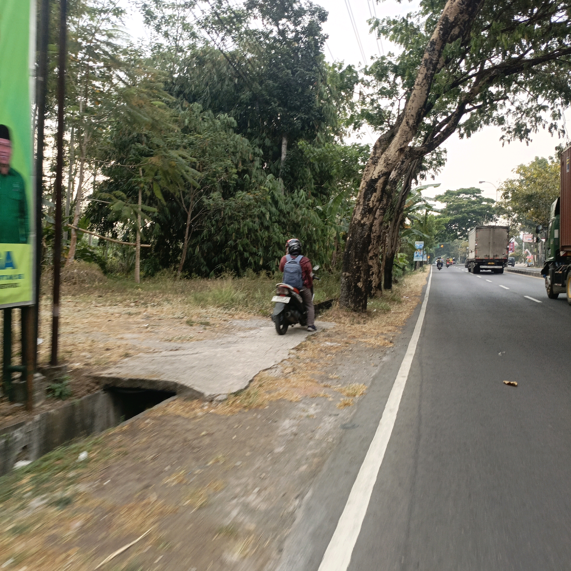 In Salatiga, Indonesia, a person rides a motorcycle with a blue backpack on a paved road. The road is bordered by a grassy shoulder and a large tree with rough, textured bark. Behind the motorcycle, a dense green forest lines the area. Further down the road, other vehicles, including a large truck, are visible. A street lamp can be seen in the distance. The sky is overcast.